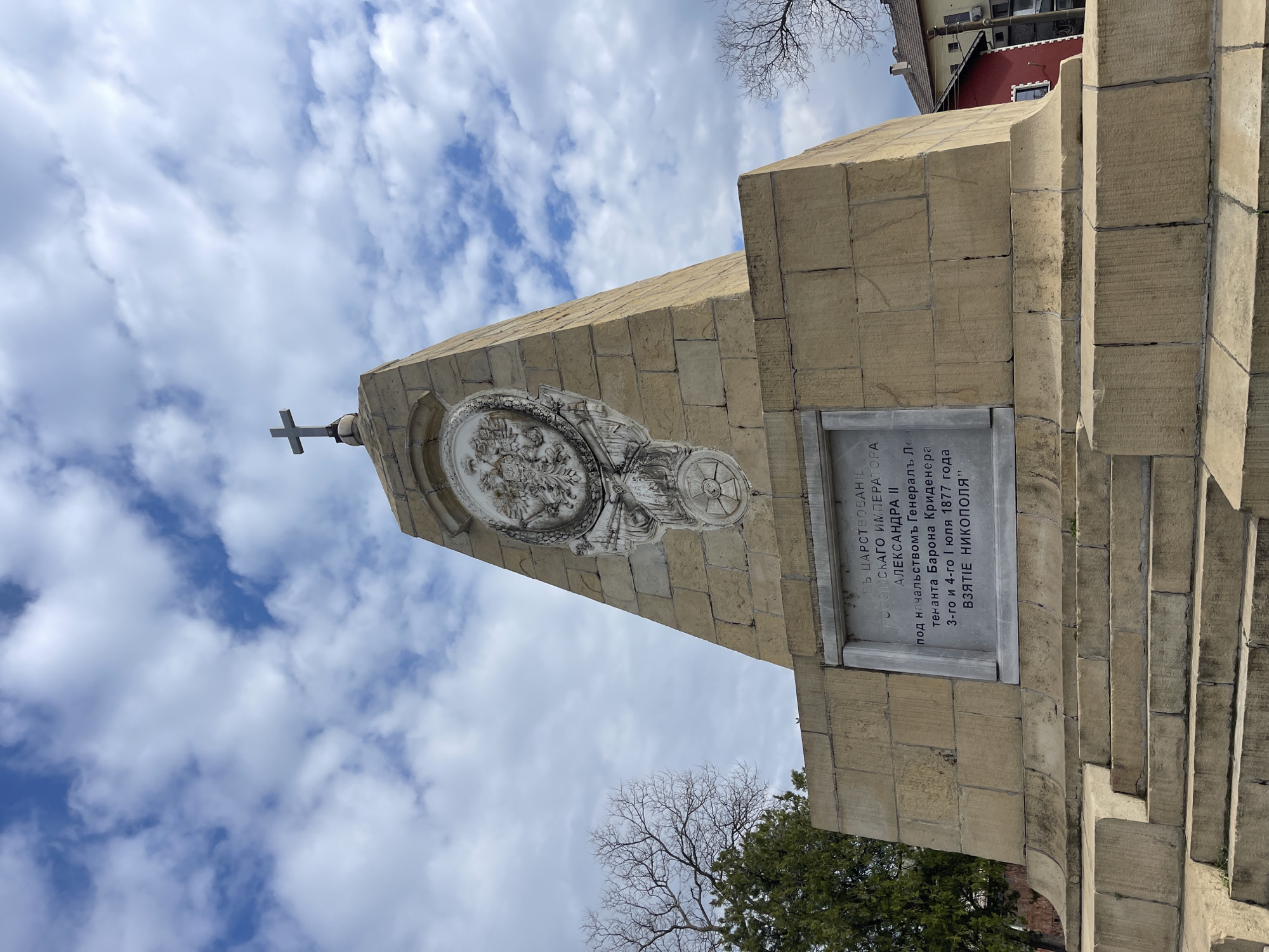 The image shows the Monument to the Liberators, specifically dedicated to the Battle of Nikopol, which took place in 1877 during the Russo-Turkish War. The monument is made of stone, featuring a cross at the top. A circular relief is placed on the monument, with a plaque below it bearing Cyrillic script. The inscription indicates that the monument commemorates the reign of Emperor Alexander II and the leadership of General Lieutenant Baron Krüdener during the capture of Nikopol on July 3rd and 4th, 1877. The sky is partially cloudy. Trees and a building are visible in the background.