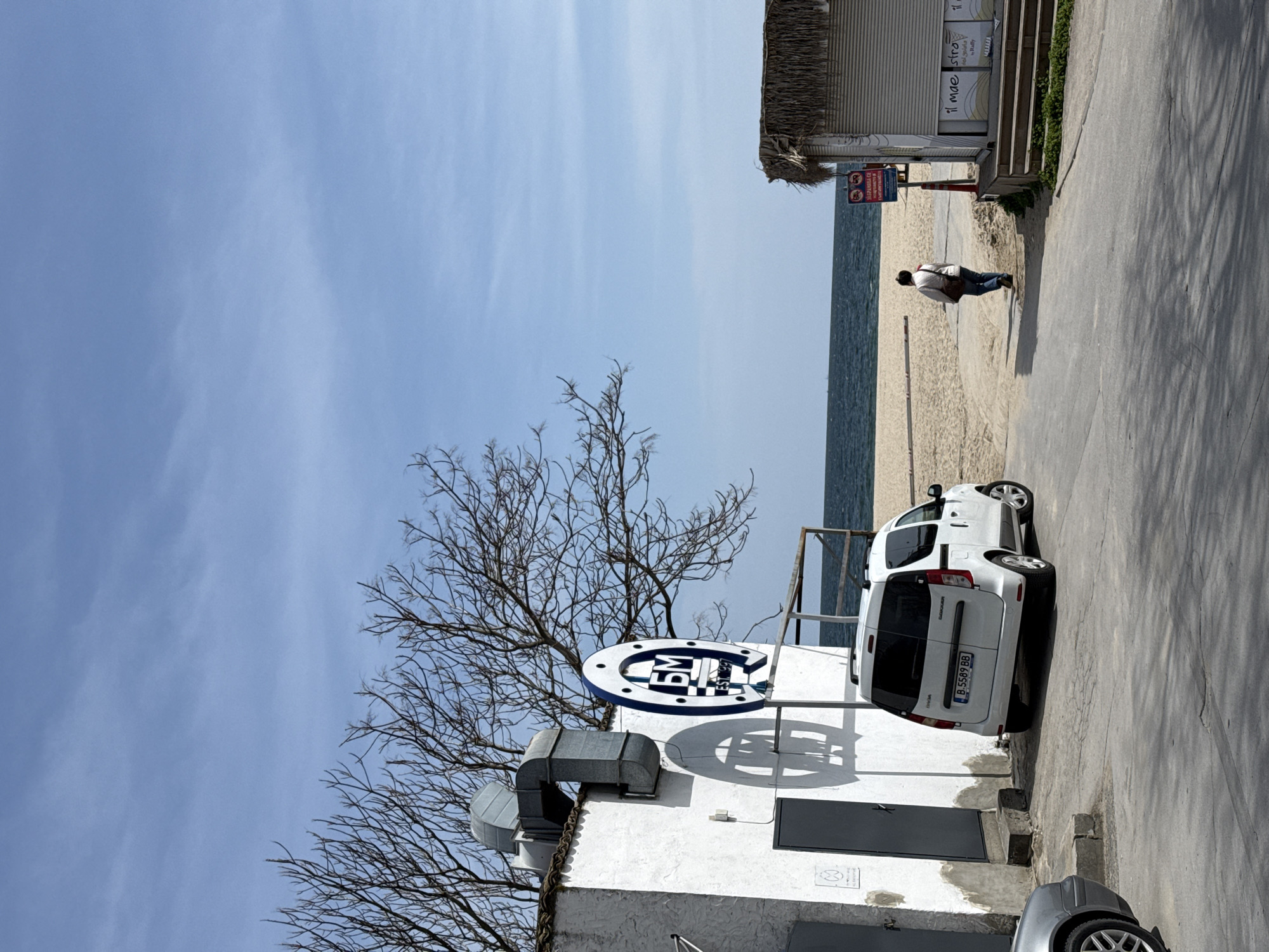 The image depicts an outdoor scene likely near a beach. A white van is parked adjacent to a white building with a sign that reads "БМ Est. 1995" with a horseshoe-like shape around the lettering. Bare tree branches are visible next to the building. A person is walking on the sandy beach towards the sea in the background. A building with a thatched roof and advertisements on its side is also visible near the beach entrance. A partial view of a silver car is seen in the bottom right corner. The scene is set under a clear, light blue sky. The scene is located in Varna, Bulgaria.