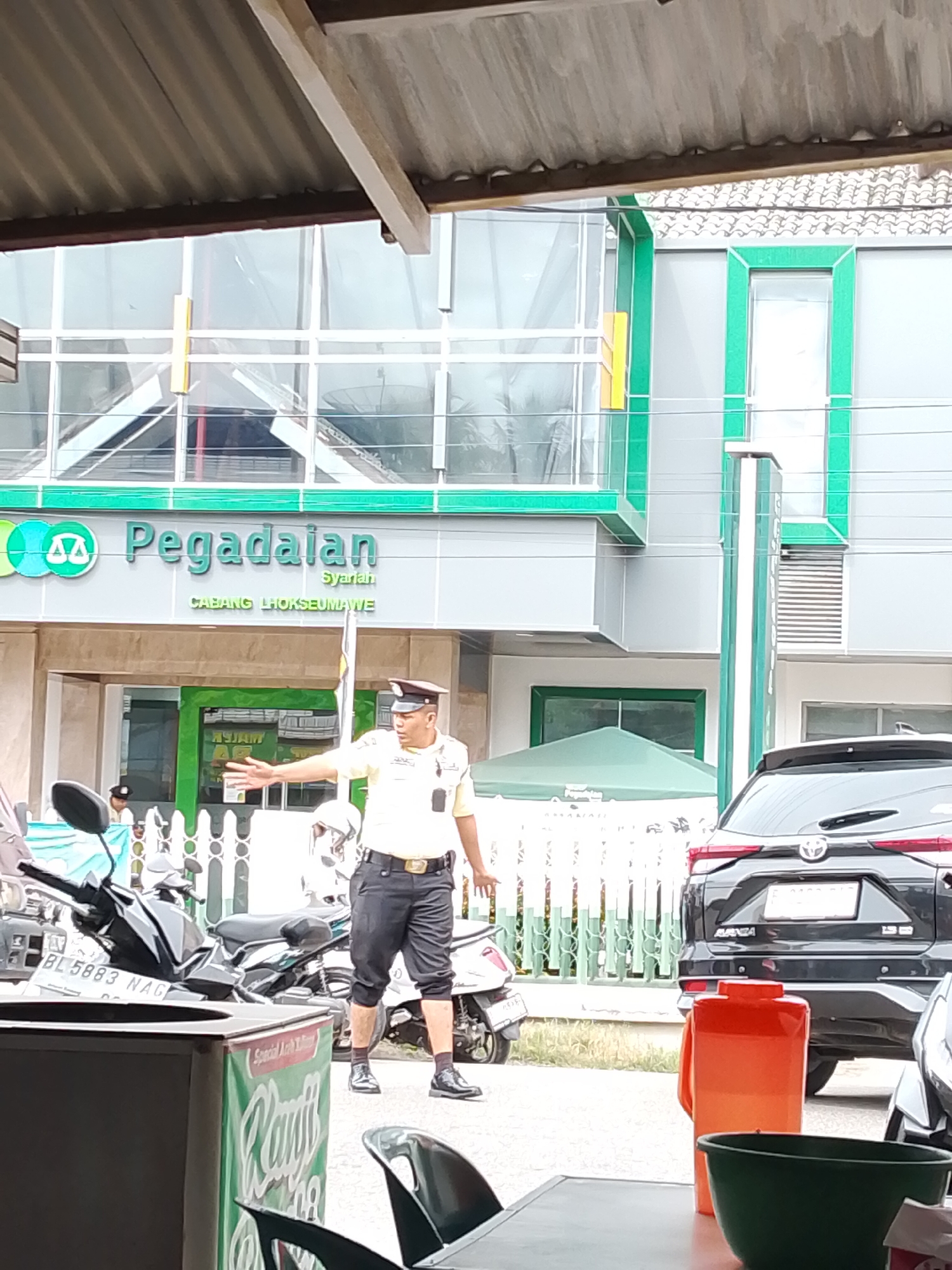 The image shows the front exterior of a Pegadaian Syariah branch in Lhokseumawe, Indonesia. A security guard in a yellow shirt and dark pants is directing traffic with his hand extended. In the foreground, there are motorcycles and a black SUV parked. The building has a modern facade with green and gray accents. The Pegadaian logo is visible on the building. The environment appears to be an urban street with paved ground.