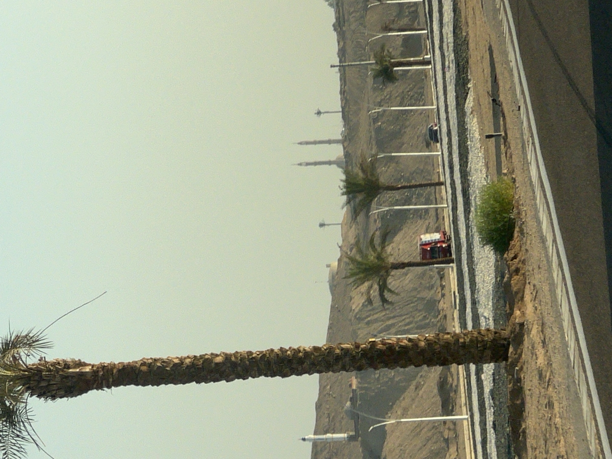 The image depicts a highway scene in Sharm El Sheikh, Egypt. In the foreground, a dark asphalt road with white lane markings is visible. To the left of the road, a tall, rough-barked palm tree stands prominently. Beyond the road, a sandy embankment rises, sparsely vegetated and featuring several modern streetlights. In the middle ground, a red truck is parked on the side of the road, and a dark-colored car is driving past. Further in the background, on top of the embankment, a white lighthouse-like structure is visible to the left, and in the far distance, several minarets rise against a hazy, pale sky, suggesting a mosque or religious complex. The overall environment appears arid and sunny, typical of the desert climate of the region.