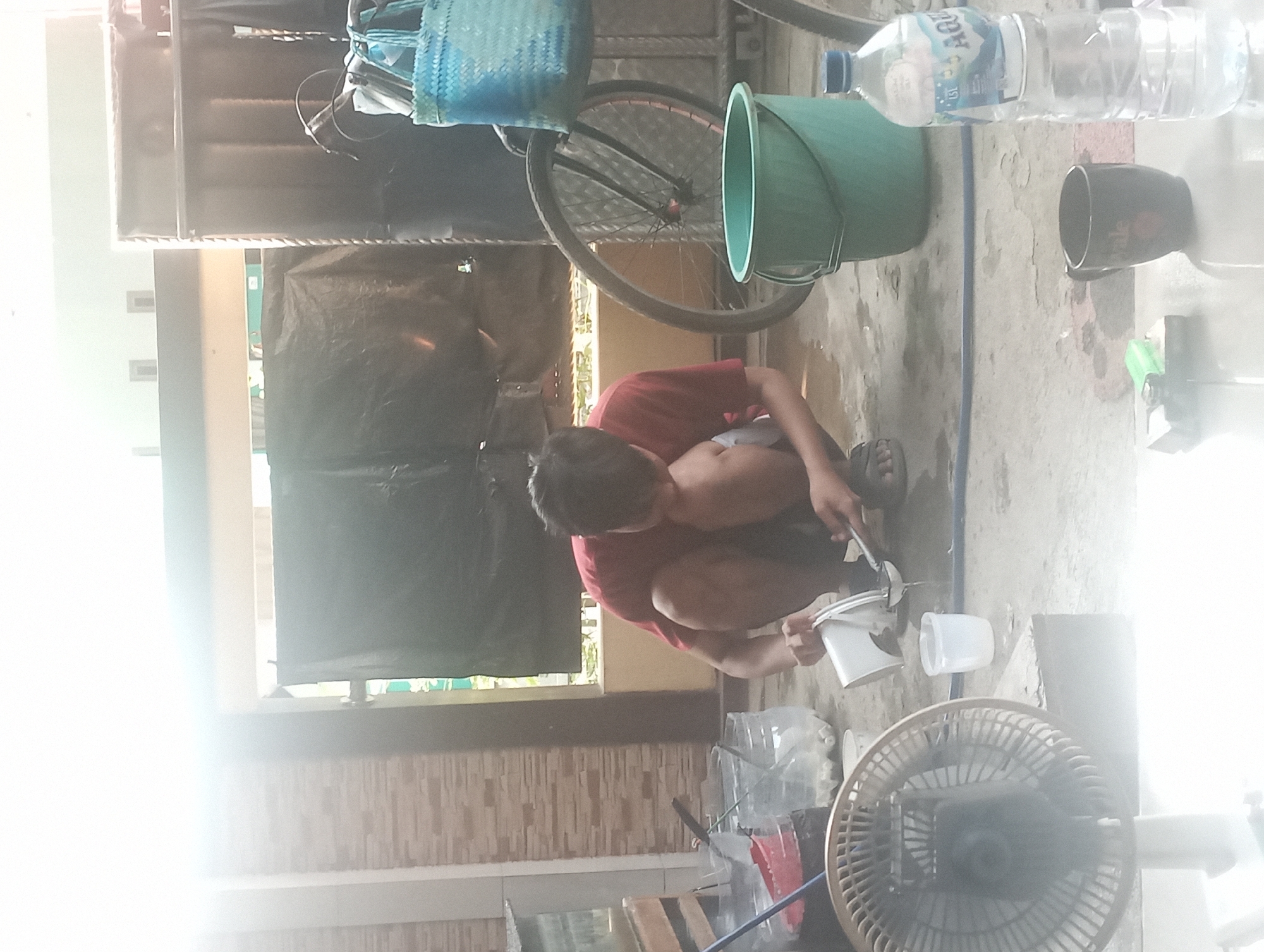 In the city of Maos Kidul, Indonesia, a young male wearing a red t-shirt is crouching on a concrete floor, engaged in the activity of transferring liquid from a white bucket into a strainer. He is positioned near several clear plastic containers filled with what appears to be gravel and water, along with a red plastic scoop. To his right, a large, teal-colored bucket stands upright, and further to the right, a bicycle with a blue woven basket attached to its handlebars is visible. In the foreground, a desk fan, a small white cup, a dark mug, a lighter, and a bottle of Aqua brand water are present. The environment suggests an outdoor or semi-outdoor setting, possibly a yard or a covered patio, with a brick wall in the background and a large black tarp obscuring a window or opening. The lighting is bright, with a strong source of light coming from the left, creating lens flare in the image.