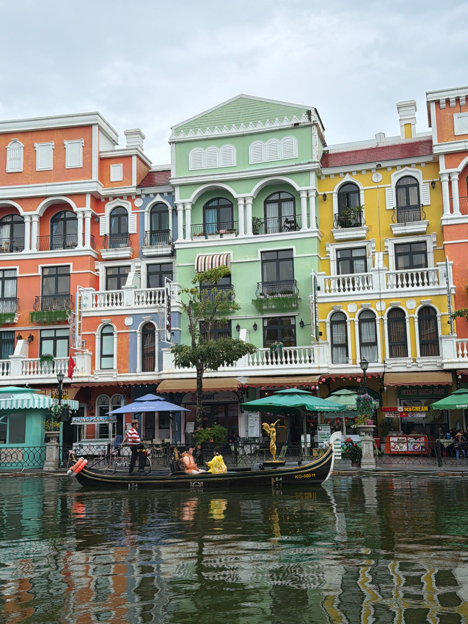 This image captures a picturesque canal scene in Phú Quốc, Vietnam, likely within a themed entertainment area designed to resemble Italian architecture. In the foreground, a black gondola navigates the calm, reflective water. A gondolier, dressed in a striped shirt and hat, stands at the stern, steering the boat. Two passengers are seated in the gondola; one wears an orange life vest, and the other is clad in a yellow raincoat. The water reflects the vibrant colors of the buildings.
Behind the canal, a row of colorful, multi-story buildings displays a style reminiscent of Venetian architecture. These buildings feature arched windows, ornate balconies, and a palette of bright hues including orange, light blue, green, and yellow. Architectural details like white columns and decorative moldings are prominent.
Along the canal's edge, there are shops and cafes, indicated by awnings, signage, and outdoor seating. Notable signs include "PHUNG PEARL HOTEL" and "SUNNY HOME STAY." Green umbrellas provide shade over some of the seating areas. A golden statue of an angel stands on a pedestal near the canal's edge, adding to the decorative ambiance. The sky above is overcast, suggesting a diffused light throughout the scene. The overall atmosphere is one of leisure and European-inspired charm.