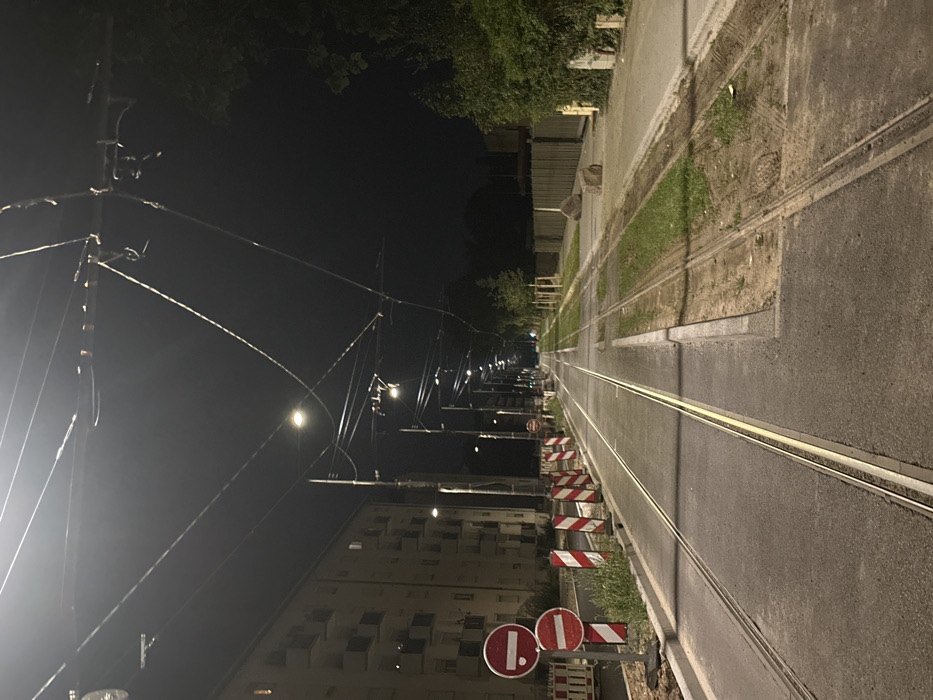 The image displays a nighttime street scene in Strasbourg, France, centered on a set of tram tracks. Two parallel tram lines extend from the foreground into the illuminated distance, marked by a series of overhead electrical wires and numerous streetlights mounted on poles. To the left, a multi-story residential building with multiple balconies stands adjacent to the tracks. In the immediate left foreground, a construction zone is delineated by two vertically stacked red and white "No Entry" signs and a row of red and white striped warning barriers. To the right of the tracks, a paved path abuts a narrow grassy median where several small trees are planted. Beyond this median, a dark fence or wall separates the public right-of-way from dense, unlit vegetation. Two large, irregular concrete blocks are positioned on the grassy median. No individuals or vehicles are visible in the scene, which is brightly illuminated by artificial light against a dark sky.
