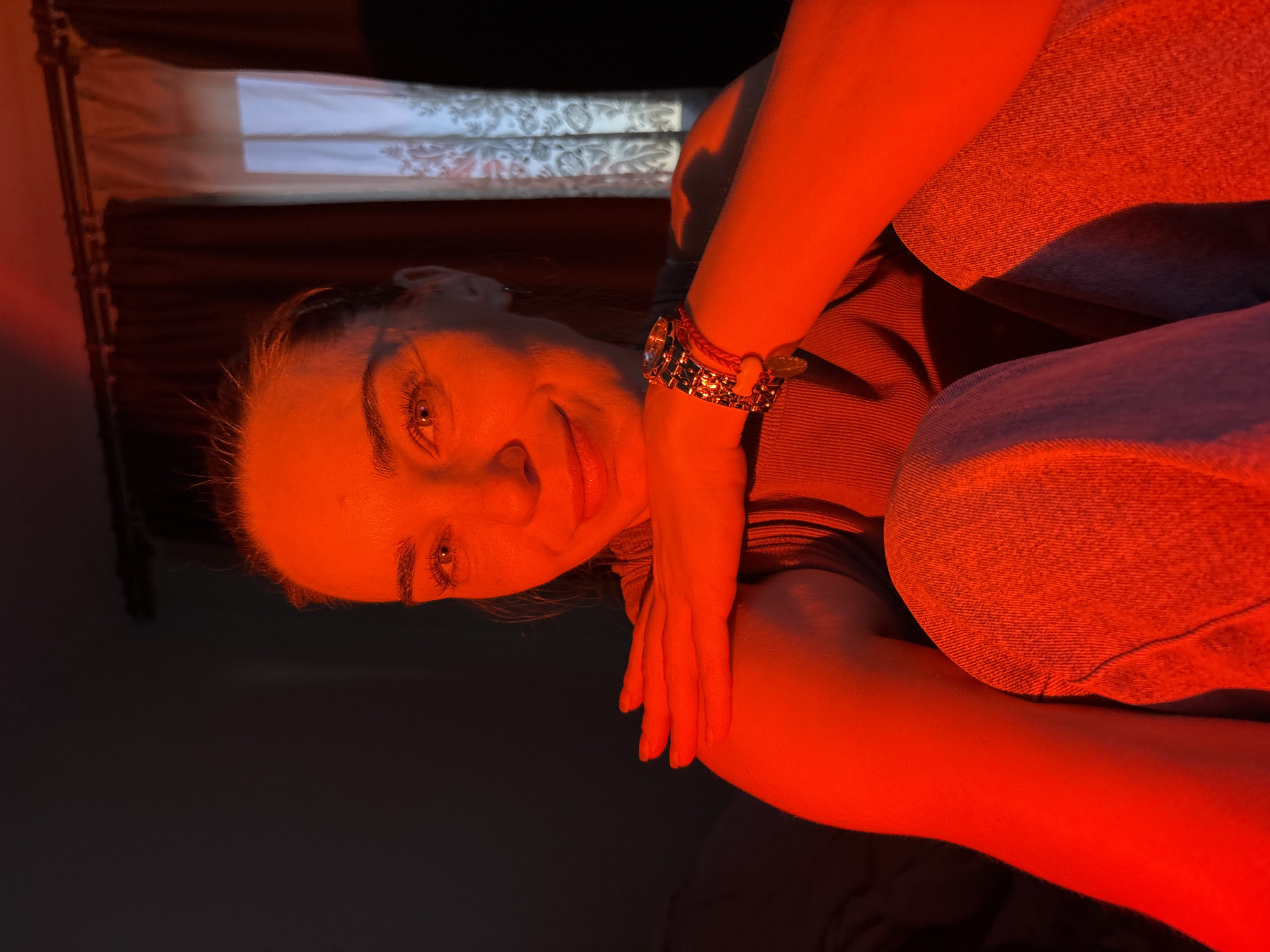 A medium shot captures a young woman in dim lighting, bathed in red light. She is sitting, facing forward, with her right arm resting on her left shoulder. Her left hand is placed on her upper arm. She is wearing a dark-colored top and light-wash jeans. A gold-colored watch with a black band is visible on her right wrist. The background is dark, with a curtain and a window visible to the right. The scene appears to be indoors, possibly a bedroom. The woman has fair skin and dark hair pulled back from her face. Her expression is neutral, with a slight smile. The red lighting creates strong shadows and highlights, obscuring some details of the environment.