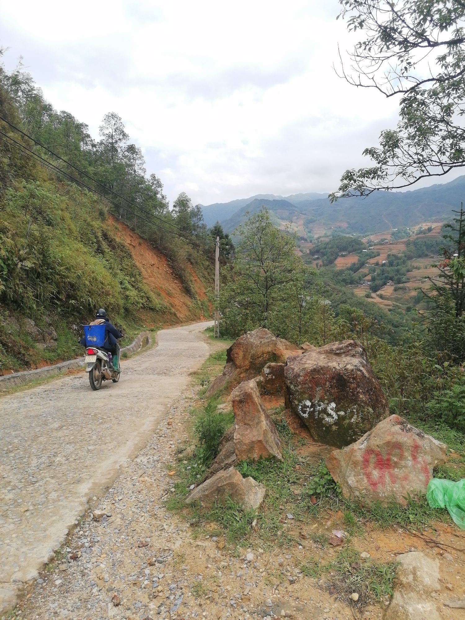 The image shows a rural road in Cat Cat Village, Vietnam. A person on a motorbike is driving away from the camera on a paved road that curves to the left. The person has a blue container strapped to the back of the motorbike. The road is bordered by a steep hill on the left, covered with vegetation, and on the right by large rocks and grass. In the distance, a valley stretches out with terraced fields and houses visible on the hillsides under a cloudy sky. Power lines run along the side of the road. A green plastic bag is on the ground near some of the rocks. The rocks are marked with white and red paint.