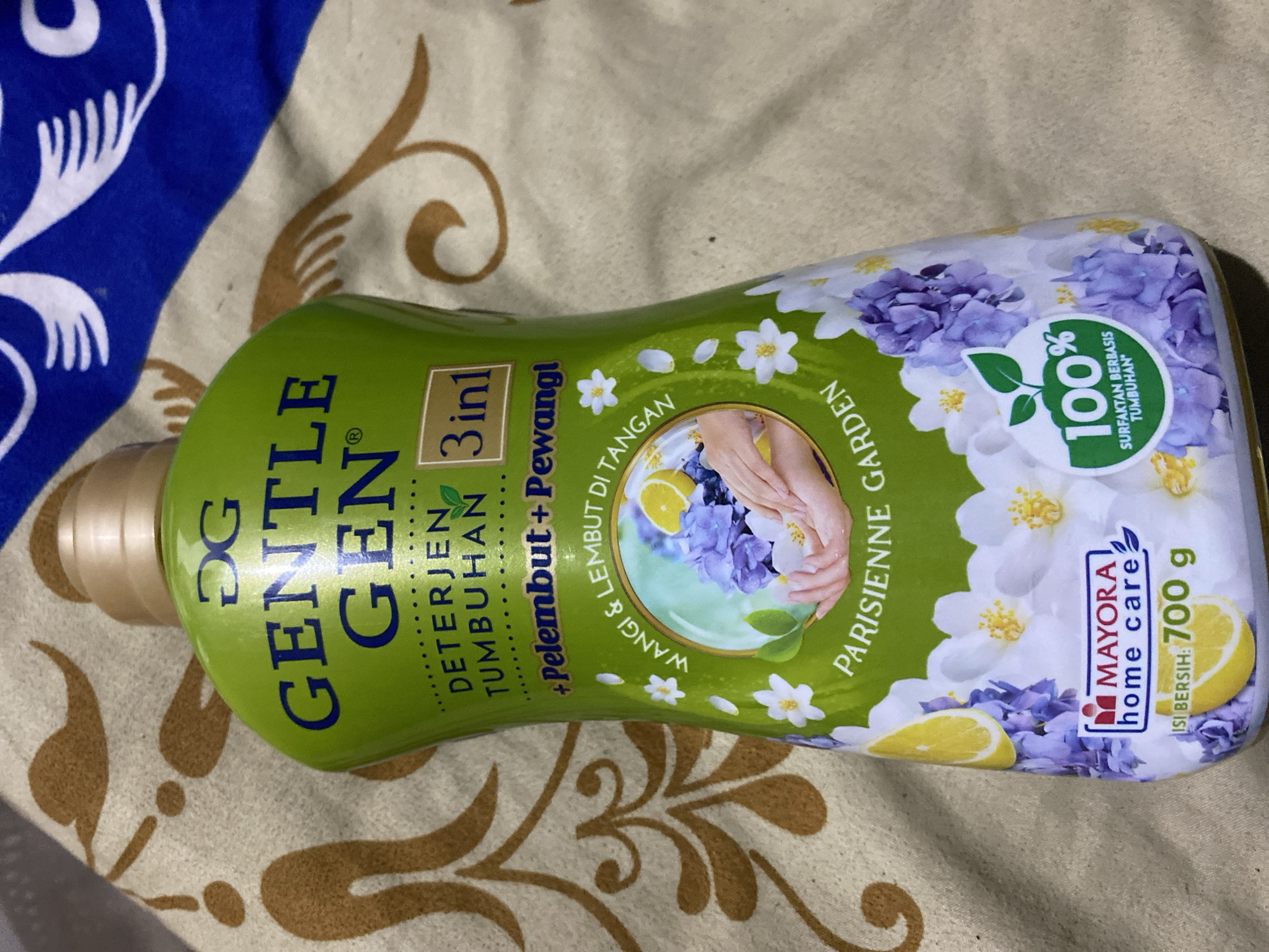 The image shows a bottle of "GENTLE GEN" laundry detergent with a green label featuring blue text and floral designs. The bottle is placed on a patterned fabric, likely a blanket or bedspread, with brown and white swirls and abstract shapes. The detergent bottle itself has text indicating it is a "DETERJEN TUMBUHAN" (plant-based detergent) and "3 in 1" functionality, including "PELEMBUT + PEWANGI" (softener + fragrance). A circular graphic on the bottle depicts hands washing with soap bubbles, lemons, and purple and white flowers. The brand "MAYORA HOME CARE" is visible at the bottom, along with the net weight "700 g". A circular green logo with "100% SURFAKTAN BERBASIS TUMBUHAN" (100% plant-based surfactant) is also present. The background fabric has a visible texture. The lighting suggests an indoor setting, possibly a bedroom or living area. There are no people visible in the image, and no identifiable landmarks that would pinpoint a specific location within Ta', Indonesia.
