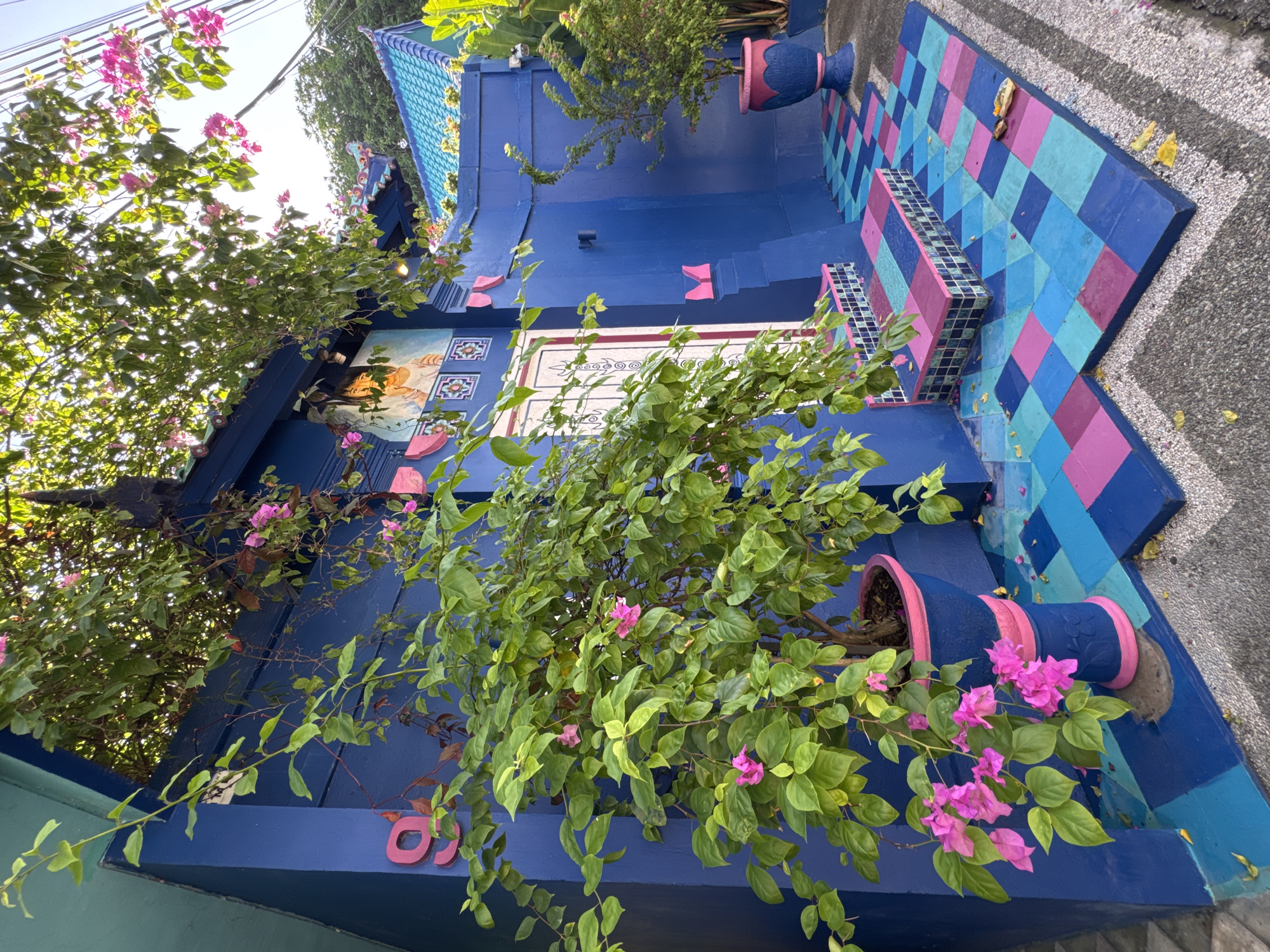 The image captures an exterior view of a vibrant, blue-painted building in Kuta, Indonesia. The structure features a tiled roof with blue and white designs, and its walls are adorned with colorful, geometric patterns, including pink and turquoise. A large bougainvillea plant in a blue pot with pink trim spills over the lower portion of the building, its green leaves and pink flowers adding to the scene's colorful aesthetic.
The entryway of the building leads to stairs painted in a checkerboard pattern of blue and pink. A smaller potted plant sits on the steps. The building's walls have a decorative panel with artwork. Part of a blue facade is visible on the left edge. The image is taken outdoors, with a clear sky and sunlight illuminating the scene. The overall impression is of a creatively decorated and eye-catching building.