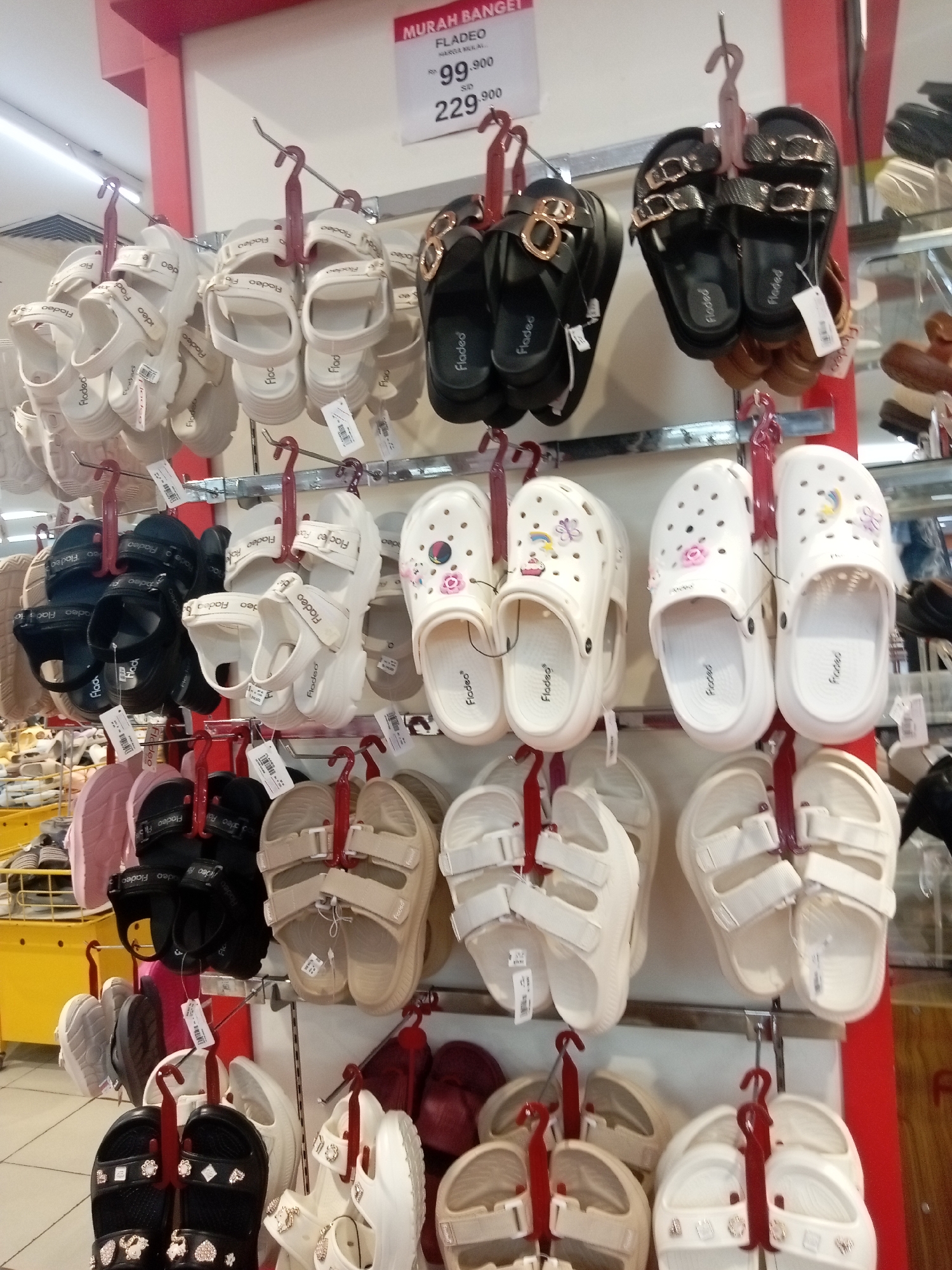 The image shows a retail display of various types of footwear, primarily sandals and clogs, in a store. The items are neatly arranged on hanging racks and shelves. Some of the footwear is white, black, or a neutral beige color. Several pairs are decorated with charms or embellishments, while others have a more minimalist design. Price tags are visible on some of the items. In the background, other merchandise and store fixtures can be seen, including shelves and part of a yellow display unit. The store appears to be well-lit and organized. The items are branded "Fladeo" and there is a sign with "MURAH BANGET" which translates to "Very Cheap". This type of merchandise and pricing suggests a retail environment, likely a shoe store or a department store section. Batam, Indonesia is a city known for its shopping malls and retail outlets. Given the context of the image, this scene is likely from a shopping mall in Batam.