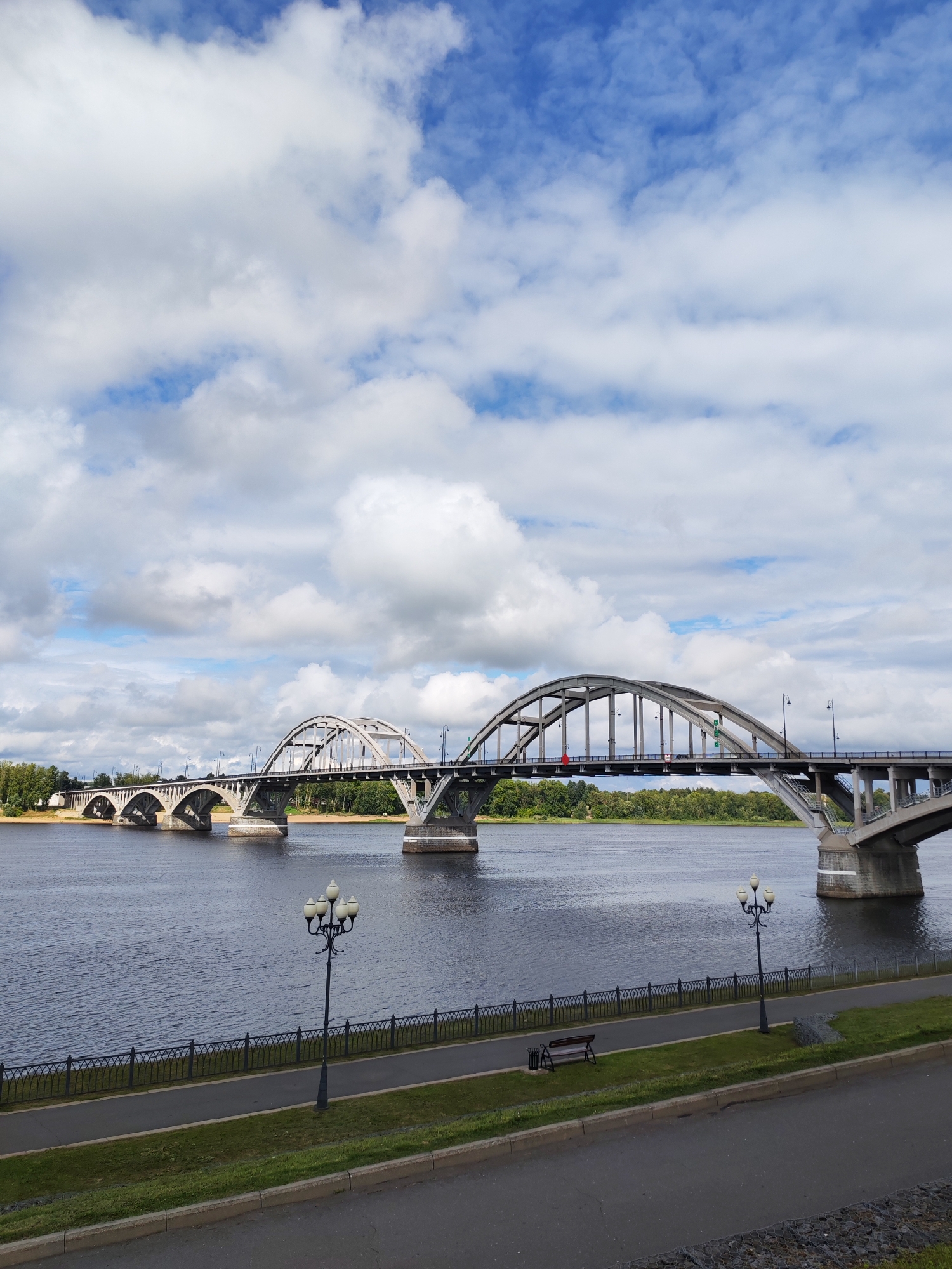 The image depicts a wide river with a multi-arched concrete bridge spanning across it. The bridge has a complex structure with several distinct sections and supporting pillars. On the far bank, a dense line of green trees is visible. In the foreground, a paved pathway runs alongside the river, separated by a decorative metal fence. Several ornate lampposts are positioned along this pathway, and a park bench is also visible. The sky above is a mix of blue with scattered white clouds. The scene appears to be a public area, possibly a park or embankment, within the city of Bol'shie Mkhi, Russia, given the architectural style of the bridge and the presence of a pedestrian walkway and lampposts often found in such locations. No people are present in the immediate foreground, but the presence of a bench suggests a place for public recreation. The water of the river is dark and appears calm.