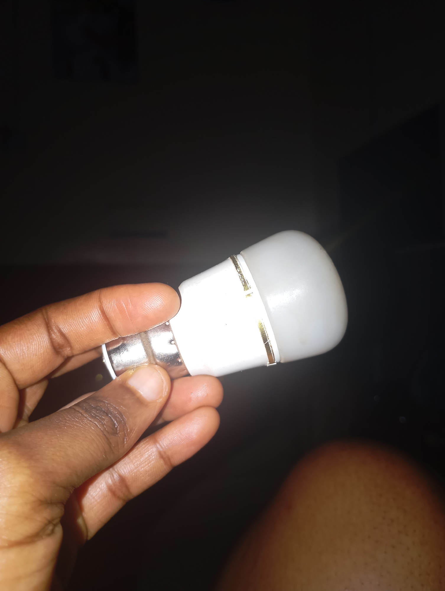 In New Stadium, Nigeria, a close-up shot shows a person's hand holding a white LED light bulb. The bulb has a metallic base and two thin gold bands around its white plastic casing. The background is dark and out of focus, with a suggestion of a person's leg in the lower right corner. The lighting is dim, with the bulb being the primary source of illumination.
