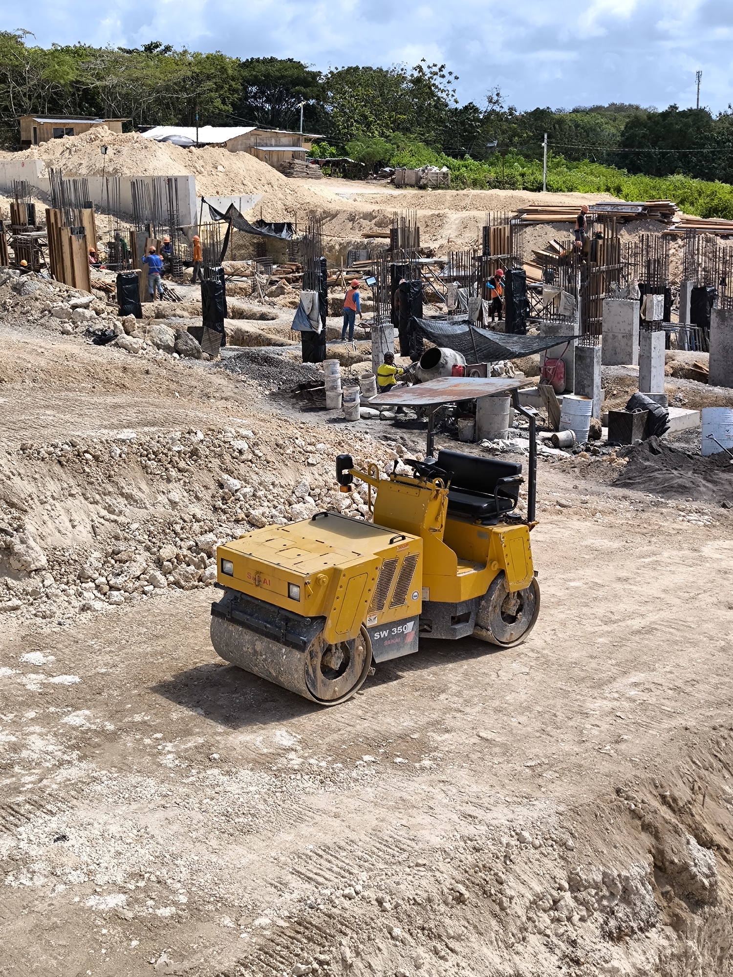 The image shows a construction site on a partly cloudy day in an unknown city. A yellow Sakai SW350 double drum vibratory roller sits on a dirt and gravel surface. In the background, workers wearing orange safety vests are engaged in various tasks around concrete pillars reinforced with rebar. The environment consists of dirt mounds, construction materials, and sparse vegetation in the distance.