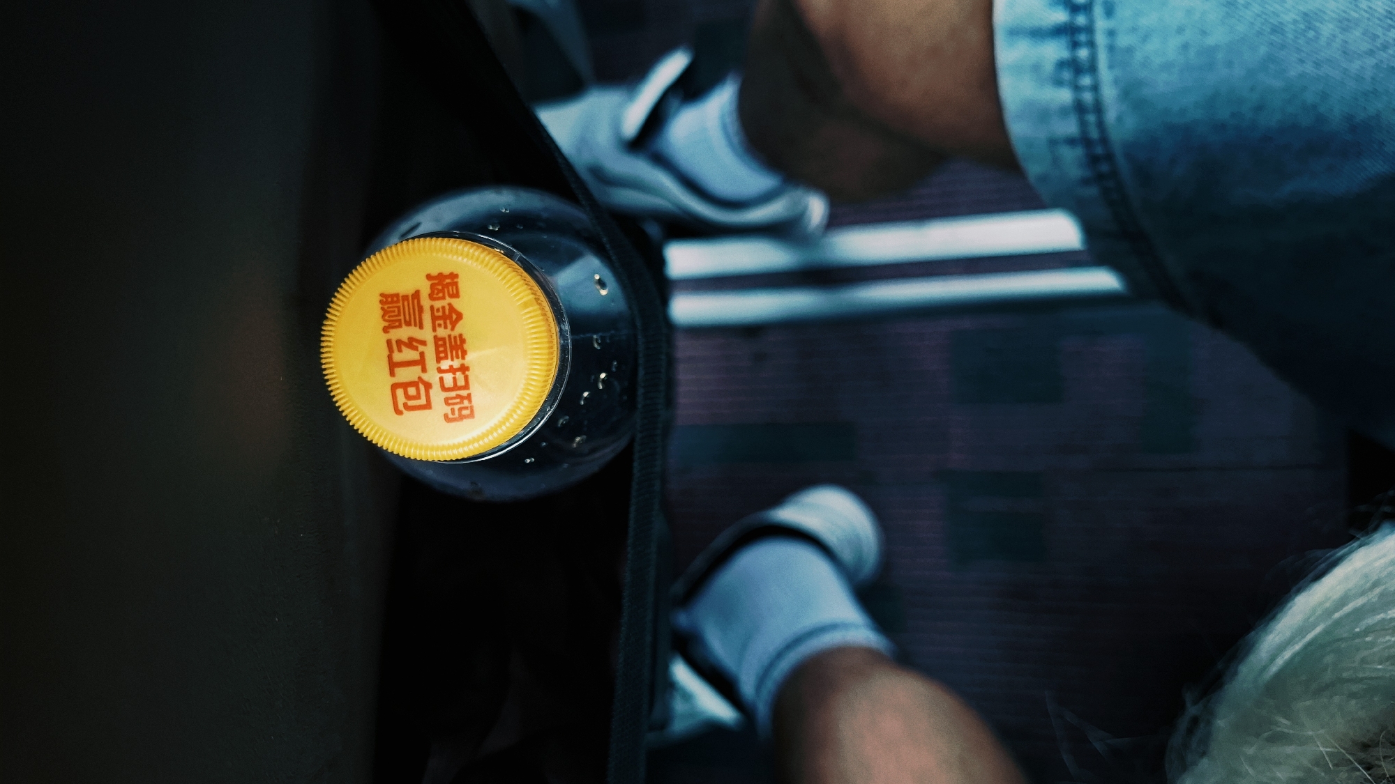 A top-down, high-angle shot shows a person sitting on a bus or train in Dongcheng District, China, with a bottle of tea and a yellow cap at their side. The subject wears light blue denim shorts, white sneakers, and grey socks. A black bag strap is across the bottle. The bottle is mostly out of focus. The background is dark with blue hues. The floor has a ridged pattern. The subject's hand can be seen in the bottom right of the image. The bottle cap has Chinese writing on it.