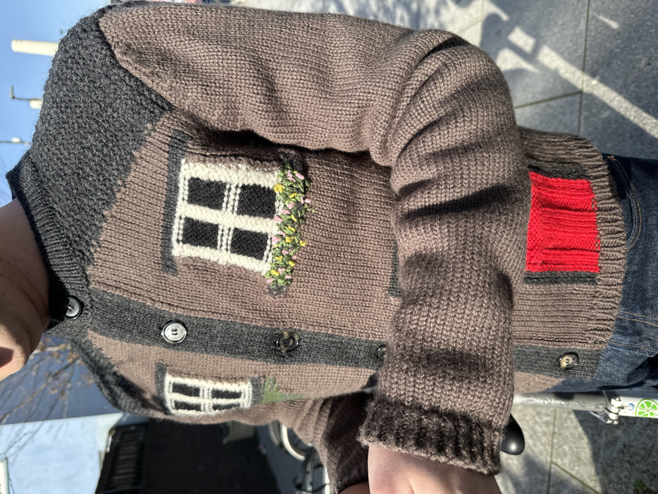 An individual stands outdoors in London, United Kingdom, on a bright, sunny day. The image captures their upper torso and arms, focusing on a unique knitted cardigan.
The cardigan is brown and dark grey, designed to resemble a house facade. Dark grey, textured sections on the shoulders mimic a roofline. Two prominent knitted windows, white with black cross-panes and white outlines, are positioned on the chest. Below the right window, an embroidered window box displays green foliage with small yellow and pink flowers. A central vertical dark grey stripe runs down the front, secured by four dark, round buttons. A rectangular, knitted red patch is located near the bottom right hem of the cardigan. The sleeves are ribbed, with the left cuff visibly rolled. Dark blue denim jeans are visible below the cardigan.
The person's hands are positioned in front, partially obscured. The immediate environment includes a paved surface. In the background, a light-colored building wall is visible to the left. A Lime electric scooter is partially visible at the bottom left. The distant background, under a clear blue sky, distinctly shows the silhouette of Battersea Power Station's chimneys, confirming the location within London. Bright sunlight casts sharp shadows across the scene.