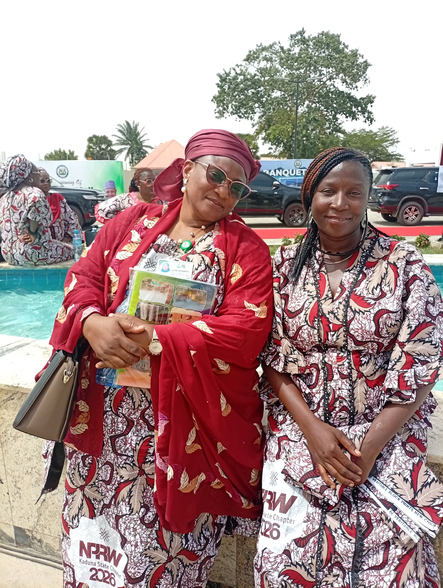 Two women are seated outdoors under bright daylight, likely at an event in Unguwan Sanusi, Nigeria. Both are smiling and looking at the camera.
The woman on the left wears a flowing red top with patterned sleeves and a matching headscarf, along with a patterned skirt. She has sunglasses on her head and a watch on her left wrist. She holds a light brown handbag and several brochures or pamphlets, one displaying text "MAUWE MOHAMMED" and images of buildings. A white patch on her skirt reads "NPRW Kaduna State Chapter 2026."
The woman on the right wears a V-neck dress made of the same patterned fabric as the first woman's skirt, featuring red, white, and brown tones. Her hair is styled in dark braids with hints of reddish-brown. She holds a folded item, possibly a piece of fabric or paper, which has text "VERITABLE BLOCK PRINTS NEW CHIG" and "RW Date Chapter 26" visible on a white patch.
In the background, a turquoise-colored fountain or small pool is visible. Other attendees, also dressed in patterned attire, are seated around the fountain. Further back, black SUVs are parked, and a white banner is partially visible with text such as "sinning Of" and "BANQUET." Large trees and a clear sky indicate an outdoor gathering or banquet setting.