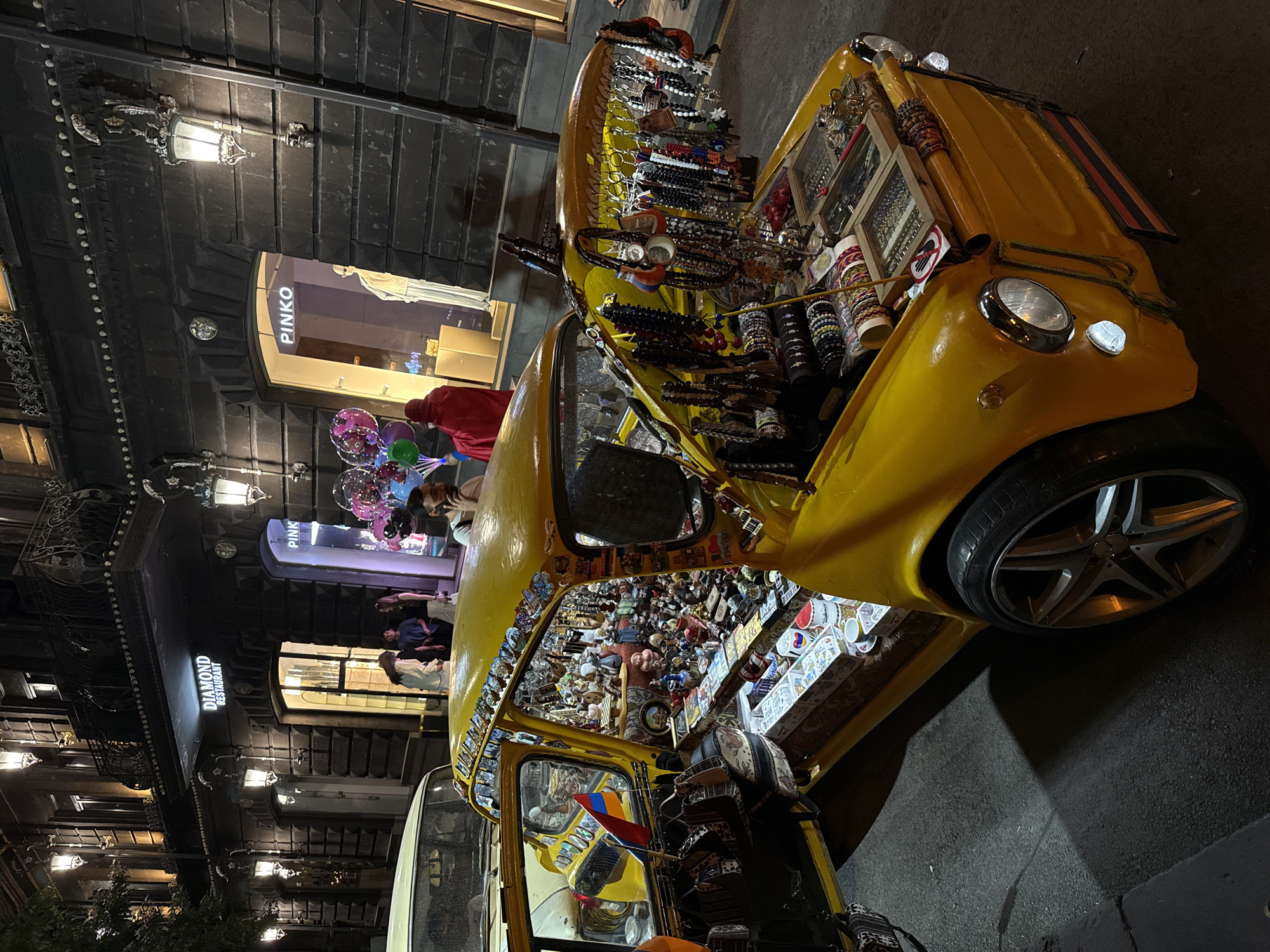 The image depicts a vibrant nighttime street scene in Yerevan, Armenia, centered around a heavily modified yellow vintage car functioning as a mobile vendor stall. The car, possibly a Zaporozhets, has its doors and front hood open, revealing a dense display of merchandise. The interior is packed with various figurines, miniature sculptures, and decorative items. An Armenian flag is visible inside the car near the dashboard. The front hood is repurposed as a display rack, holding numerous strands of necklaces, bracelets, and other beaded jewelry hanging from a custom frame, alongside trays of rings and smaller items. Small decorative plates or magnets are affixed to the car's roofline and windshield frame. The car features modern five-spoke alloy wheels and a small striped plate, mimicking the Armenian flag, attached to its front bumper.
In the background, an ornate, dark-colored stone building with illuminated wall-mounted lanterns and decorative balconies houses commercial establishments. A prominent sign for "DIAMOND RESTAURANT" is visible above an entrance, and a "PINKO" fashion store displays mannequins in its brightly lit window.
Several individuals populate the scene. To the right of the yellow car, a person is partially visible, speaking on a mobile phone. To the left, a person clad in a red hooded garment holds a cluster of luminous, colorful balloons, facing away from the viewer. Other pedestrians are visible walking or standing near the entrances of the background buildings. The street is paved with dark asphalt, and the artificial lighting casts clear shadows.