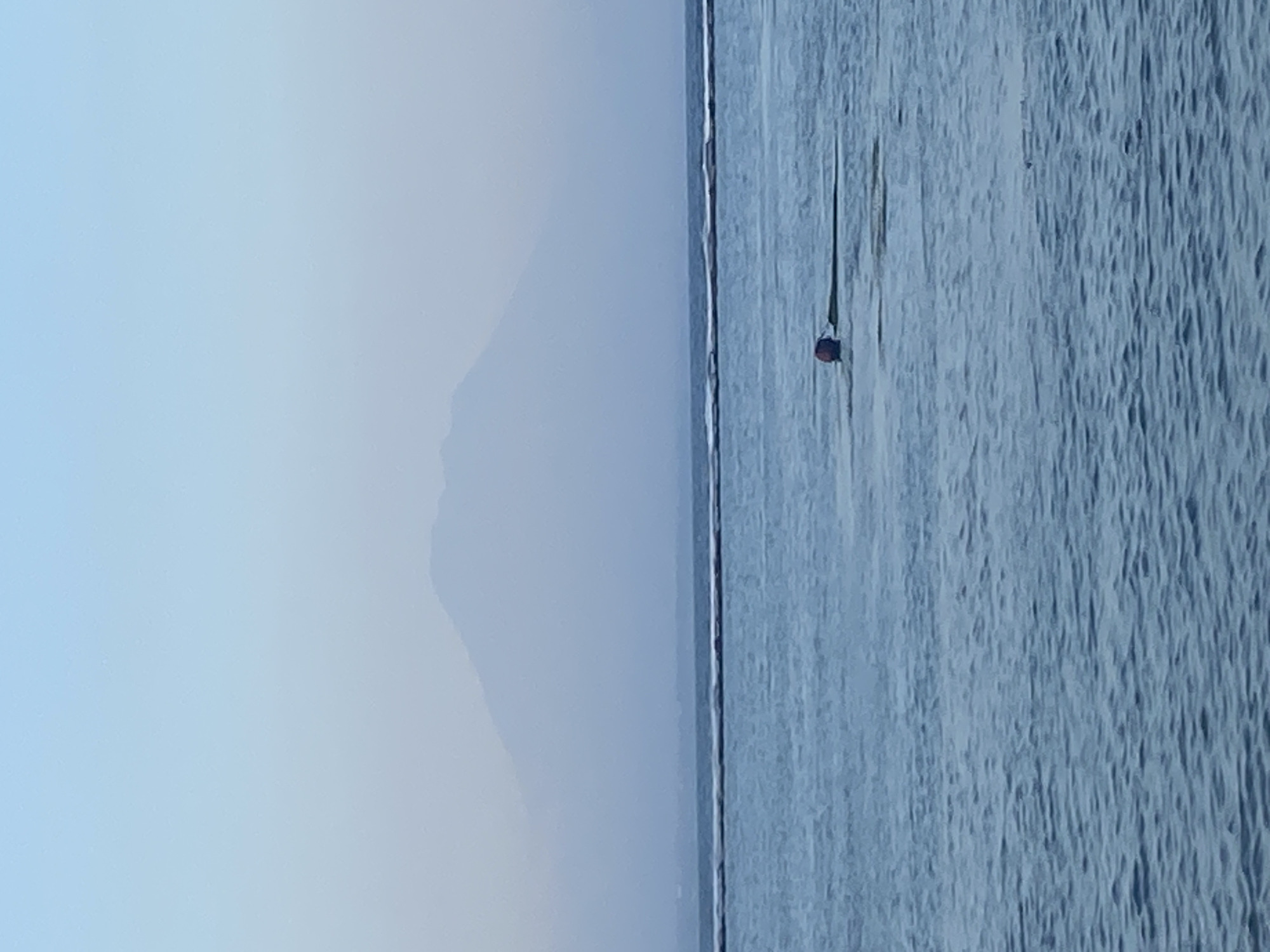 In the waters off Denpasar, Indonesia, a serene ocean scene unfolds under a pale sky. A faint mountain range, shrouded in mist, forms a distant backdrop. Closer to the foreground, a line of buoys marks a boundary in the water. A single red buoy, bobbing on the gentle waves, is the focal point of the water. The ocean's surface is a tapestry of subtle ripples, reflecting the muted light of the sky.
