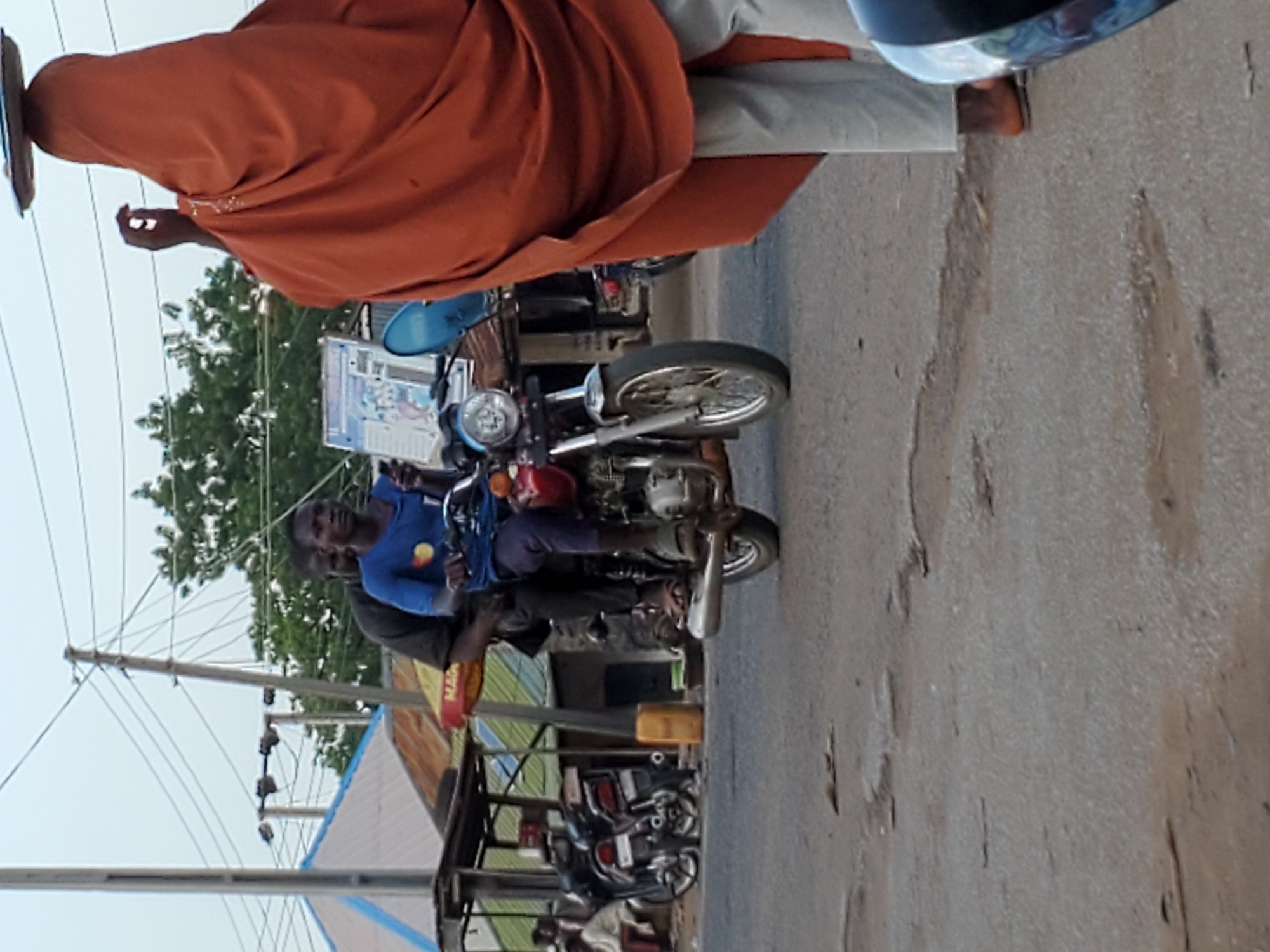 This image captures a vibrant street scene during daylight hours in Mararaba, Nigeria. The sky appears bright but somewhat hazy.
In the foreground, on the left side of the frame, a person wearing a flowing reddish-orange garment, possibly a robe or abaya, is visible from the waist up. They are seen in profile, with their right arm raised, holding a flat, rectangular object, possibly a mobile phone or small tablet.
The central focus of the image is a motorcycle. A person wearing a blue short-sleeved shirt and dark trousers is seated as the driver, looking directly towards the viewer with a neutral expression. Another individual is riding as a passenger behind them. Attached to the handlebars of the motorcycle is a rectangular sign or poster, primarily white and blue, featuring some graphics and text that are largely unreadable due to distance and angle.
Further into the background, other motorcycles are parked along the side of the road. On the right, parts of buildings with corrugated metal roofs are visible. A yellow and red object in the midground, possibly a sign or part of a vehicle, displays the partial text "MAG". Utility poles and numerous overhead wires stretch across the scene, particularly on the left. The ground is a paved road, showing some wear and tear. The overall impression is a typical bustling street in an urban or semi-urban environment.