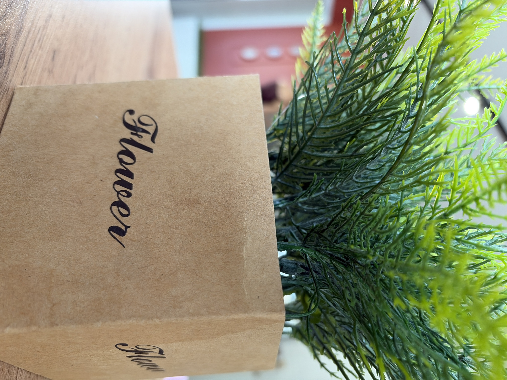 The image shows a close-up of a green artificial plant in a brown cardboard pot with the word "Flower" printed on its side in black calligraphy. The pot is on a wooden table. The background is blurred, showing hints of a room interior with a pink wall and a bright light source. There are no people in the image. The scene is indoors.