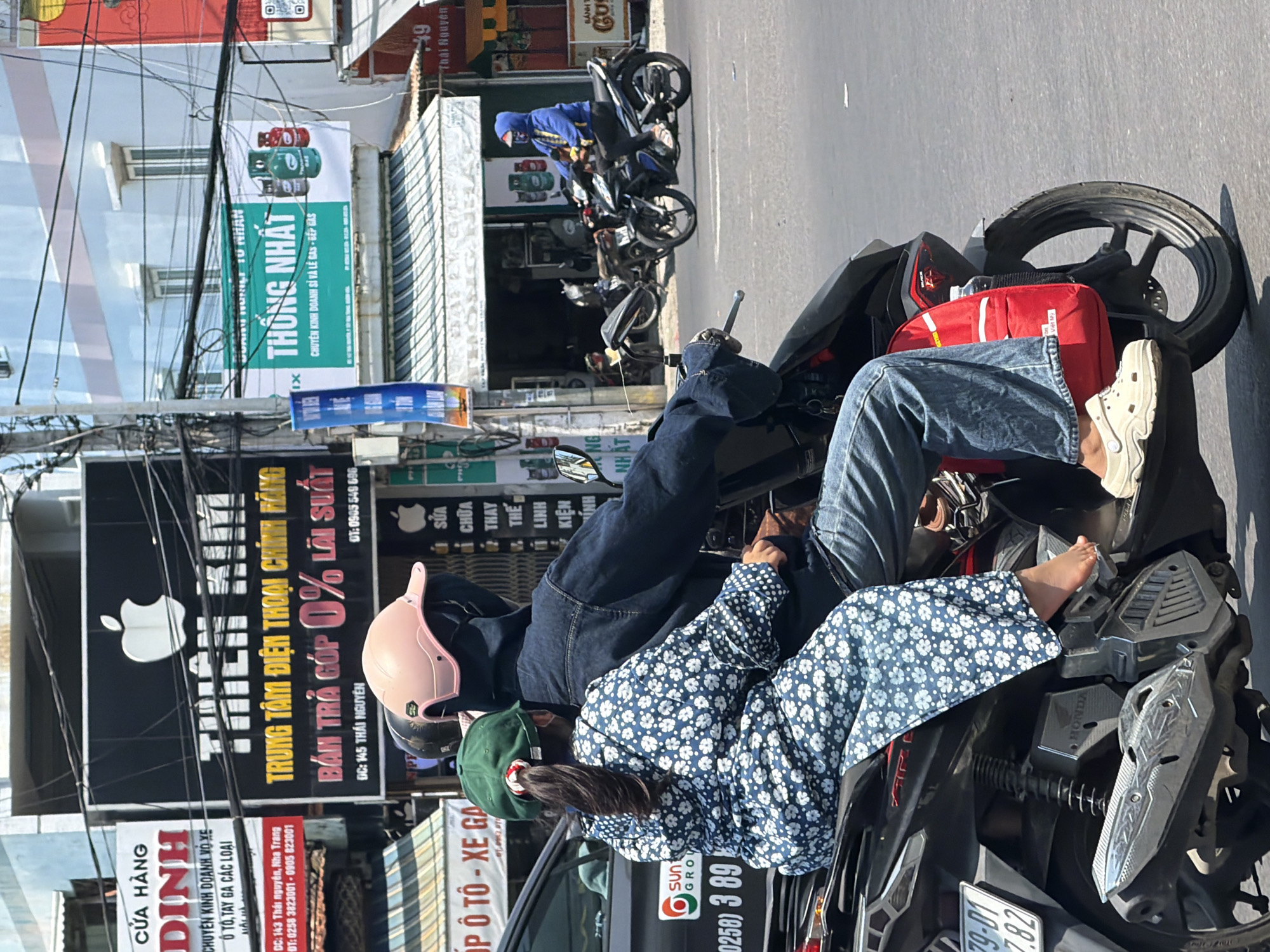 This is a close-up, eye-level outdoor photograph of two people riding on a motorcycle on a paved road in Tay Nha Trang Ward, Vietnam. The rider, wearing a dark blue hoodie, jeans, and a light pink helmet, is driving the motorcycle. Seated behind the rider is a young child wearing a blue and white floral print jacket, a green hat, and a dark face mask. The child's left foot is bare and rests on the motorcycle. A red backpack is attached to the back of the motorcycle, behind the child. In the background, buildings and storefronts are visible, including signs with Vietnamese text. One prominent sign features the Apple logo and reads "THIEN KIM". Another sign promotes "THONG NHAT" with images of gas cylinders. The scene is illuminated by bright daylight, casting distinct shadows.