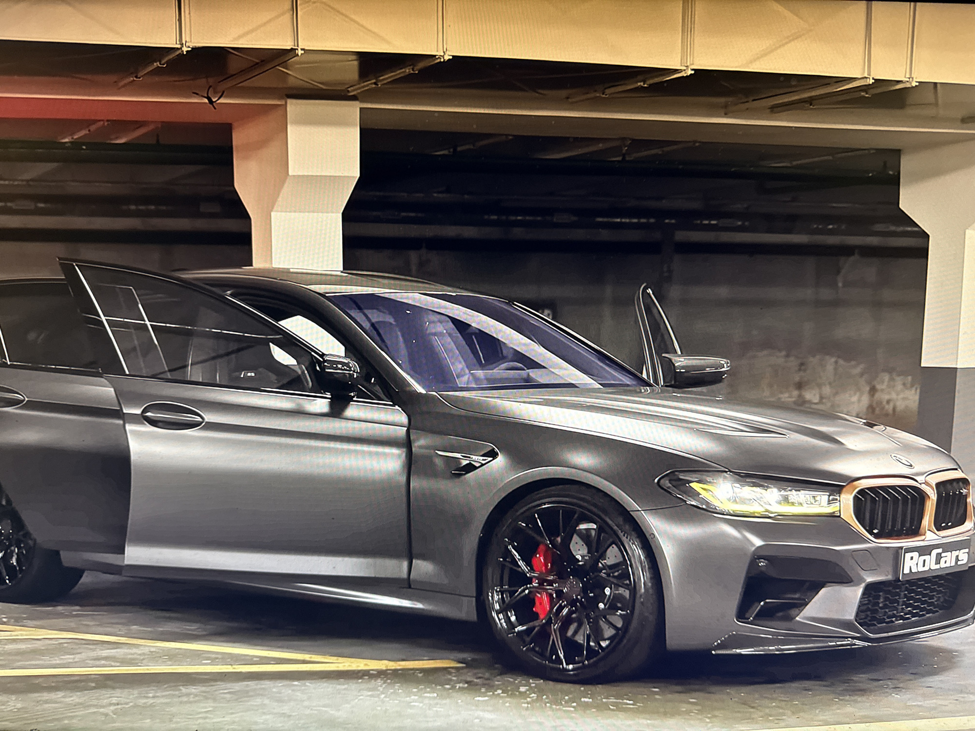 A gray BMW M5 is parked in an indoor parking garage. The car's front and rear doors are open. The car has black rims with red brake calipers. The grill is copper-colored. The word "RoCars" is visible on the front grill. Yellow parking lines are visible on the floor. The parking garage has overhead piping and ventilation systems. The city of Sofia, Bulgaria is not mentioned or visible in this image.