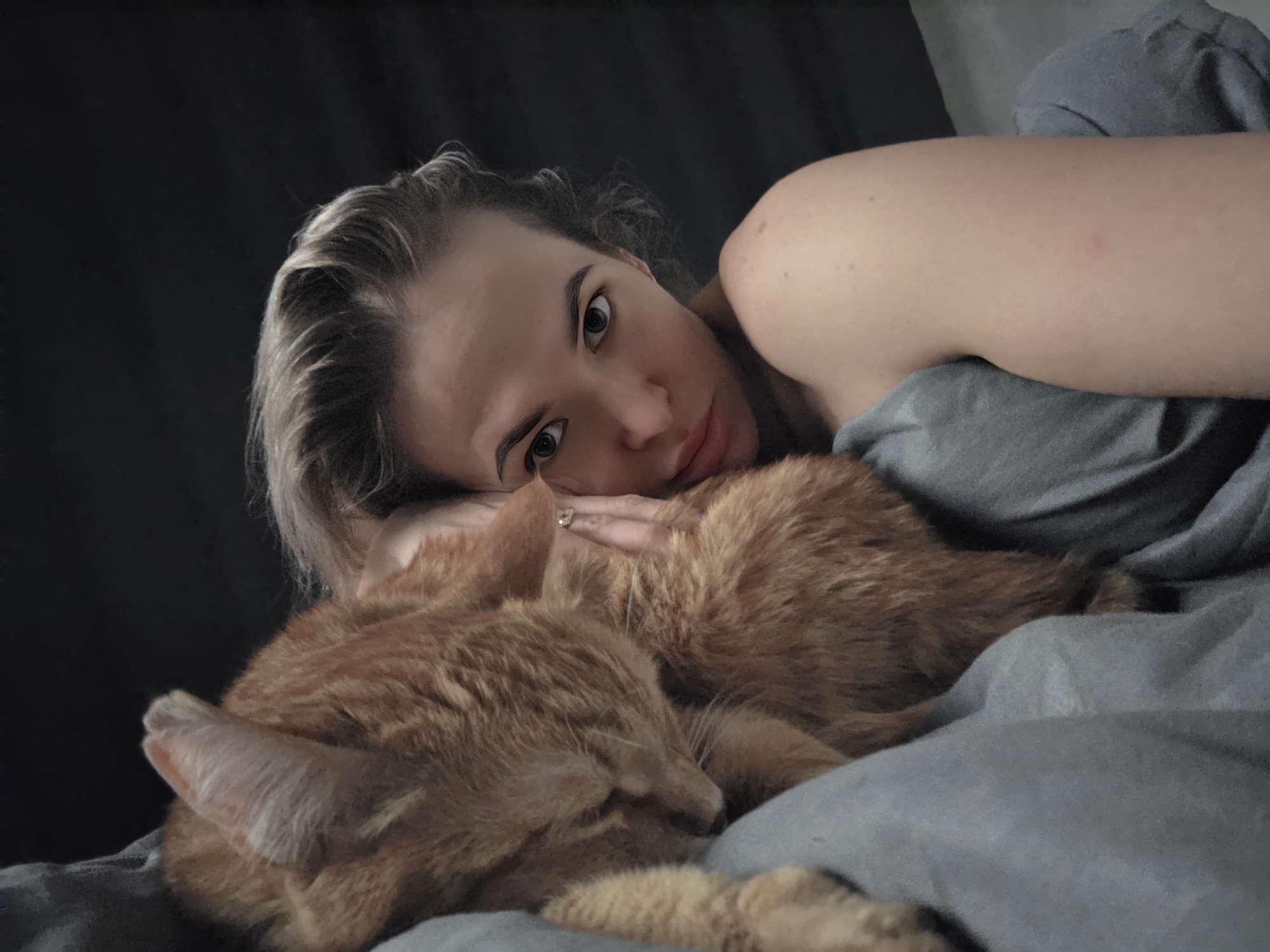 A woman is lying in bed with an orange cat. The woman is fair-skinned with blonde hair. She is wearing a ring on her right hand. The cat is curled up next to her, and both are resting on grey bedding. The location of the image is unknown, and there is no indication it was taken in Chaikovsky, Russia.