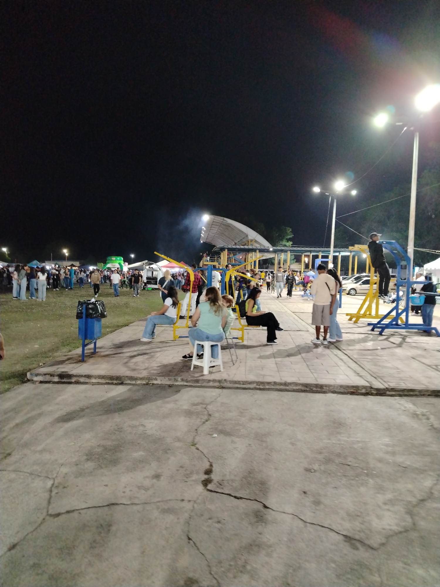 The scene depicts a public square in Tinaquillo, Venezuela at night. Overhead lighting illuminates the area, highlighting a gathering of people. Several individuals are seated on outdoor exercise equipment, while others stand nearby. A crowd of people is visible in the background, suggesting a public event. A trash can and other park fixtures are present, and the ground is cracked and worn. A structure with a curved roof is also visible in the background.