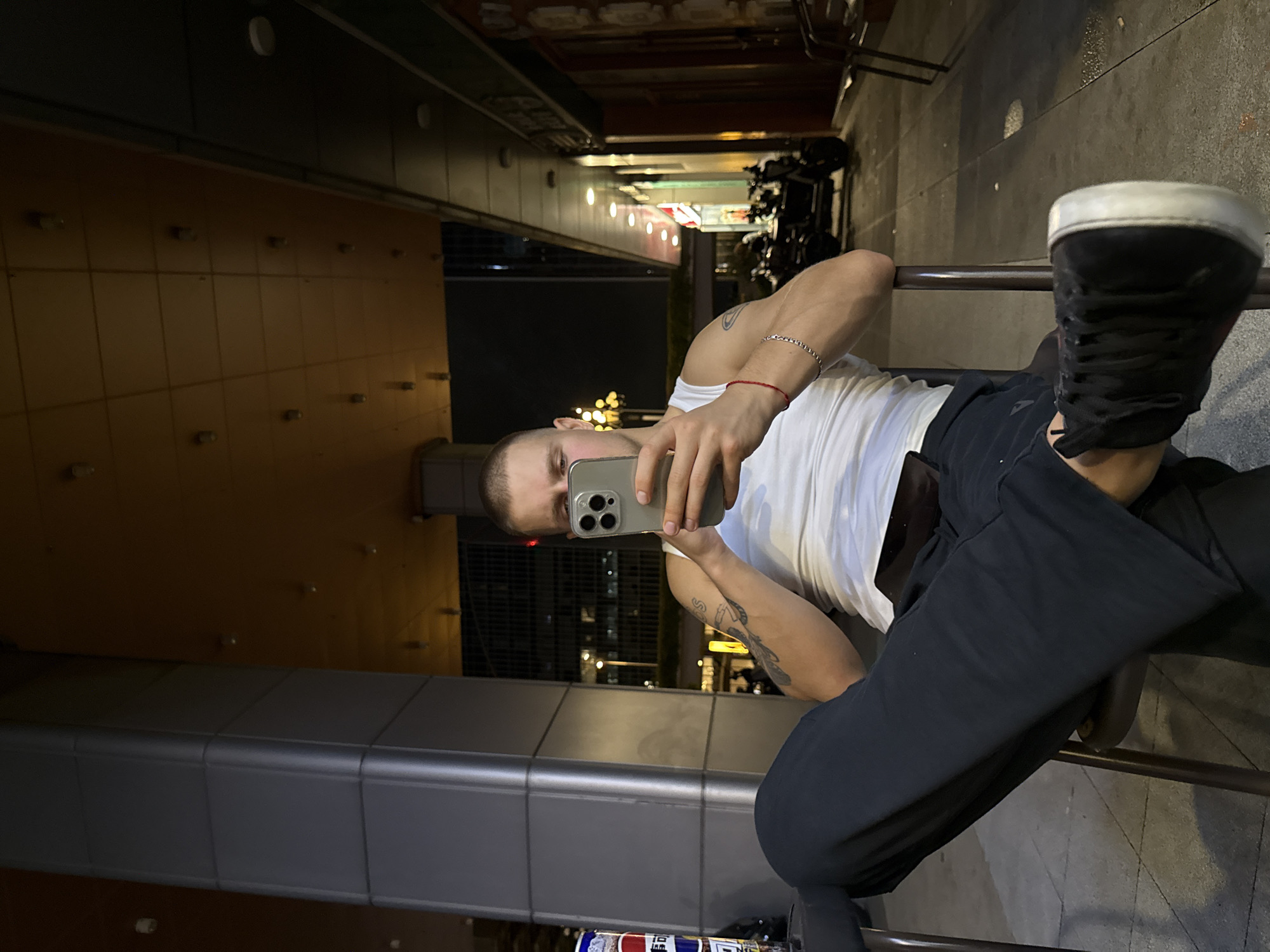 A man is seated on a chair, outdoors at night, in what appears to be a commercial district in Huangpu District, China. He is wearing a white tank top and dark pants, with black sneakers. The man is holding a smartphone and appears to be looking at it. The background shows a building with illuminated signage and several motorcycles parked outside. The lighting suggests it is nighttime, with artificial light sources illuminating the scene. The man has tattoos on his arms and is wearing bracelets.