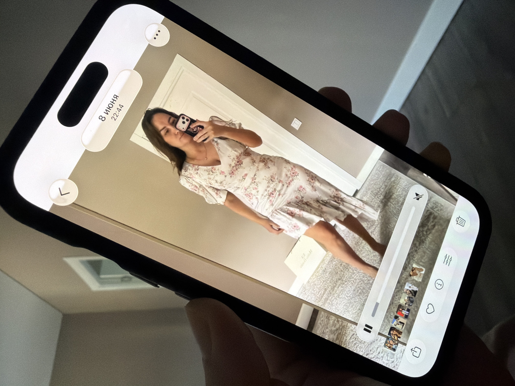 A person is holding a smartphone, displaying a mirror selfie of a woman in a floral dress. The woman is standing in a room, reflected in a mirror. The smartphone screen shows a video player interface with a progress bar, thumbnail previews, and various icons for sharing, liking, information, settings, and deletion. Above the video player, there is a timestamp indicating "8 июня 22:44" (June 8th, 10:44 PM). The background of the room appears to be a domestic interior with neutral-colored walls and flooring. The scene is set indoors. There is no discernible connection to the city of Malinovka, Russia.
