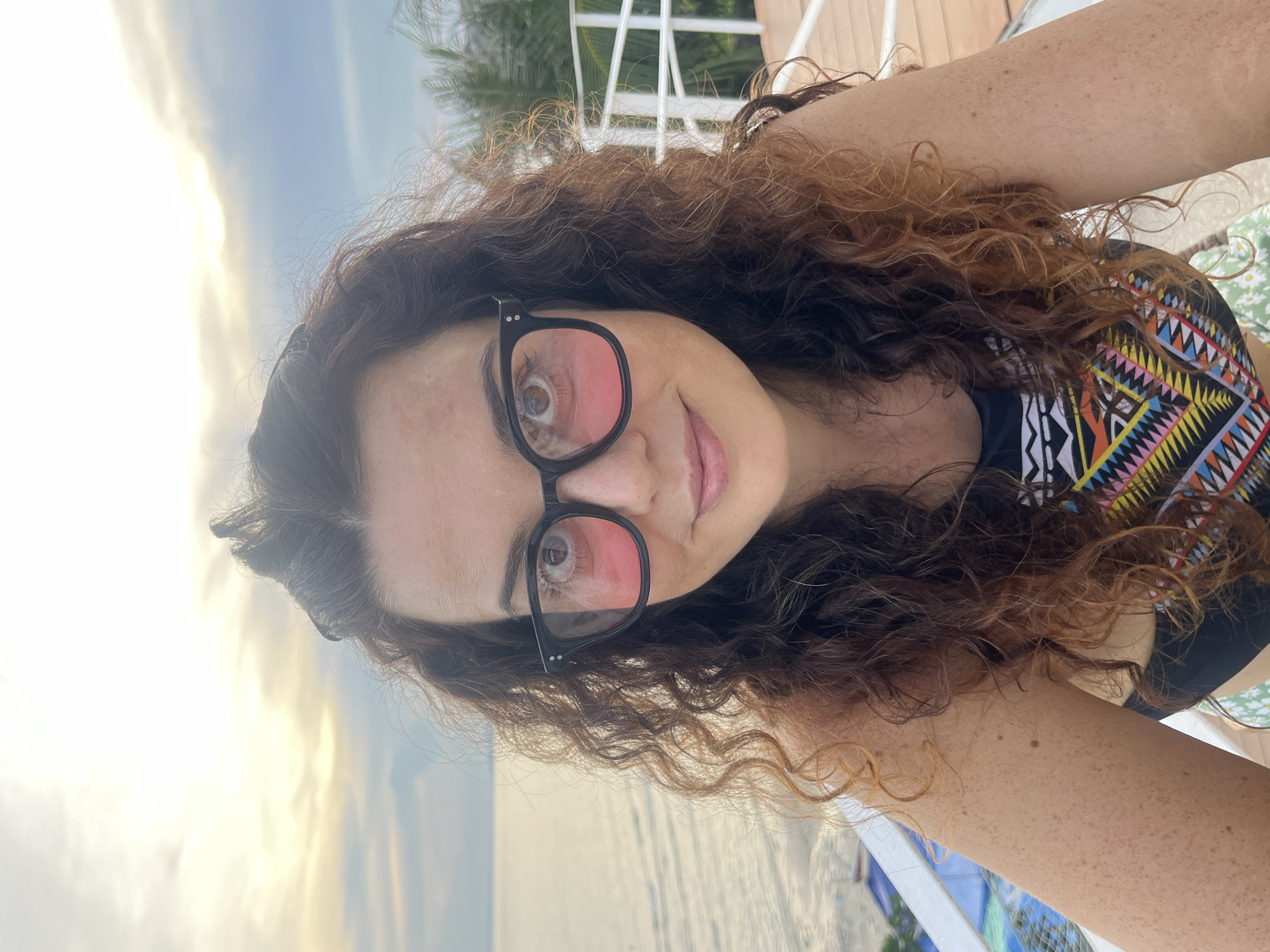 This is a close-up, selfie-style photograph of a woman with long, curly brown hair, some strands of which are lighter brown or auburn. She is wearing large, black-rimmed glasses with pink-tinted lenses that reflect the sky. Her skin has a light tan and is dotted with freckles. She appears to be wearing a black top with a colorful, patterned neckline, possibly a swimsuit or rash guard, featuring geometric designs in black, white, yellow, orange, pink, and blue. Her left arm is visible, showing freckles and some slight redness, perhaps from sun exposure.
The background suggests an outdoor, tropical setting. The sky is bright with soft, diffused light, indicating it might be early morning or late afternoon. There are hints of palm trees and a white railing, suggesting a resort, balcony, or waterfront area. The calm water of the sea is visible in the lower part of the image. The overall mood is relaxed and casual. The image was taken in Mũi Né, Vietnam. No visible text is present in the image.