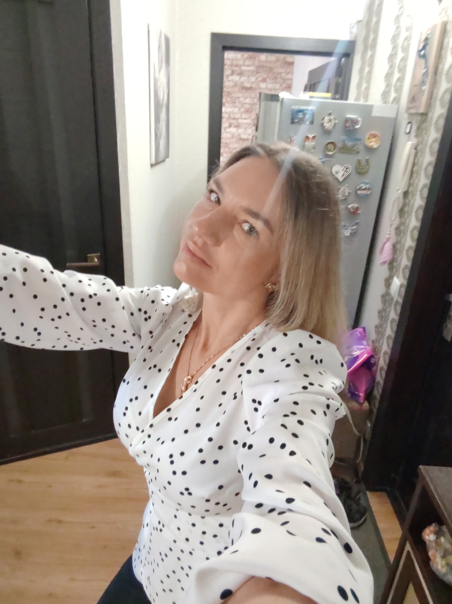 A woman is captured in a selfie, likely indoors within a home in Petropavlovsk-Kamchatsky, Russia. She appears to be of middle age, with light blonde hair that extends past her shoulders. Her light-colored eyes are directed towards the camera, and she has a neutral to slightly confident expression. She is wearing a white blouse patterned with small black polka dots, featuring a V-neckline that reveals a gold-colored necklace with a pendant. The blouse has long, slightly puffed sleeves.
The background shows an interior setting, possibly a hallway or kitchen. To the left, a dark-colored door is partially visible, and above it on the light-colored wall hangs a framed picture or artwork in monochrome tones. Prominently featured to the right of the woman is a stainless steel refrigerator, heavily adorned with a variety of magnets. These magnets display diverse designs, including an owl, hearts, a turtle, a coffee cup, and other shapes, suggesting souvenirs or personal decorations. Beyond the refrigerator, patterned wallpaper with a light base and darker wavy designs is visible, along with another dark door frame and a small dark wooden shelf unit holding decorative items. A crinkled purple object, possibly a balloon or gift wrapping, can be seen near the bottom right of the refrigerator. The floor is covered with light-colored wood laminate or parquet. No discernible text is visible on any objects in the image.