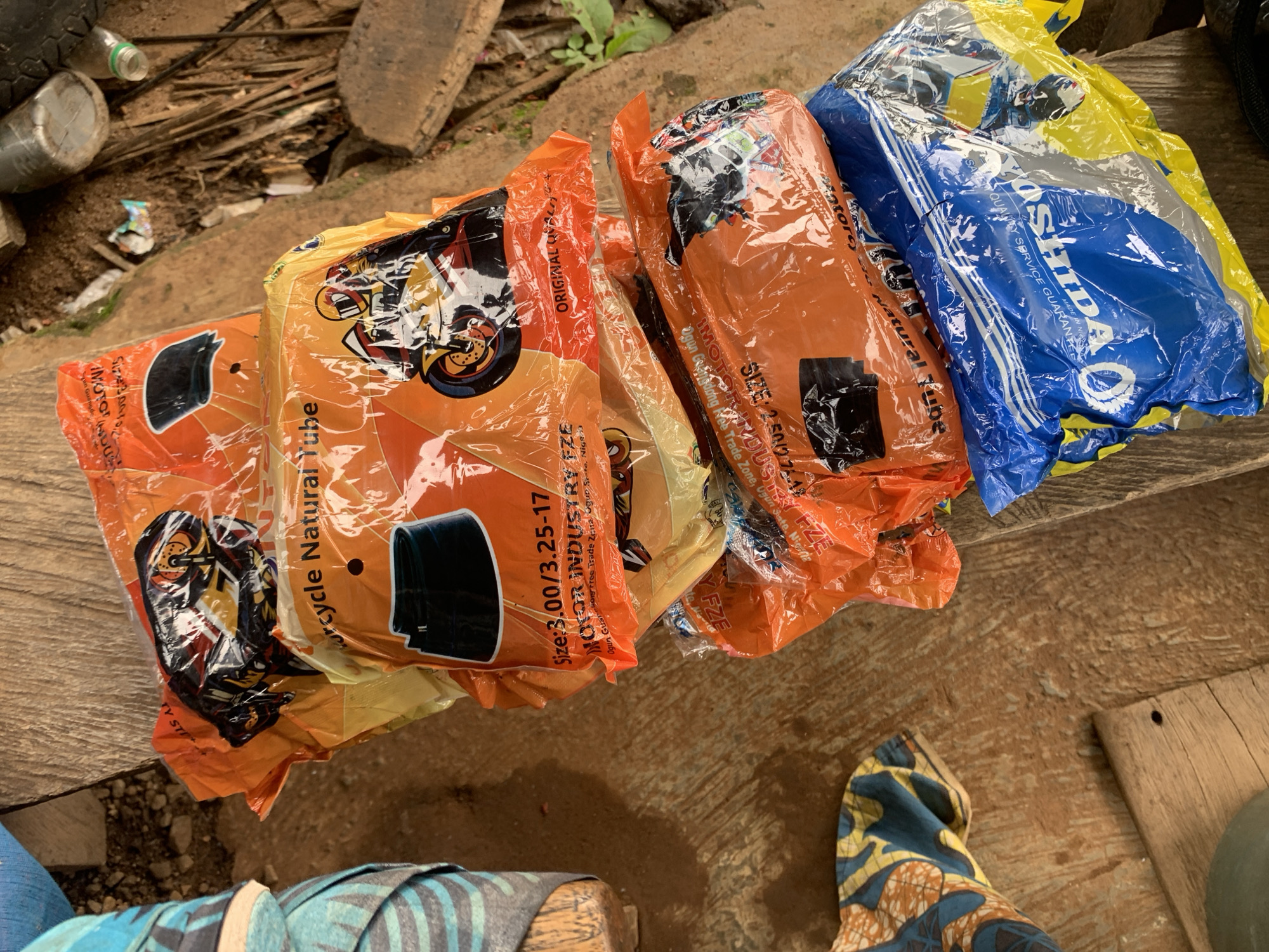 The image displays a close-up, high-angle view of several packaged items, likely motorcycle inner tubes, stacked on a rough wooden surface. The majority of the packages are made of clear orange plastic, revealing the contents. Each orange package features an illustration of a motocross rider on a motorcycle. Visible text on these orange packages includes "Motorcycle Natural Tube", "SIZE: 3.00/3.25-17", and "SIZE: 2.50/2.75-17". Further text mentions "MOTOR INDUSTRY FZE", "ORIGINAL QUALITY", and details like "Ogun Guangzhou Free Trade Zone, Ogun State, Nigeria", providing a specific location cue.
To the right, one package is distinctive in blue and yellow plastic, with the brand name "YOSHIDA" clearly visible, along with phrases like "PREMIUM QUALITY SERVICE GUARANTEE" and "MADE IN NIC".
The setting appears to be outdoors during daytime, indicated by the bright, natural lighting. The wooden surface beneath the packages is rough, and the background shows a cluttered, earthy area with debris, including a dark old tire, plastic bottles, and small pieces of wood, suggesting an informal outdoor environment. Parts of patterned fabric items, possibly clothing or bags, are visible in the bottom corners of the frame. The presence of the packages, particularly with the "Ogun State, Nigeria" text, implies a commercial or storage context, possibly in a market or roadside stall in Gbeleko, Nigeria. No people are visible in the frame.