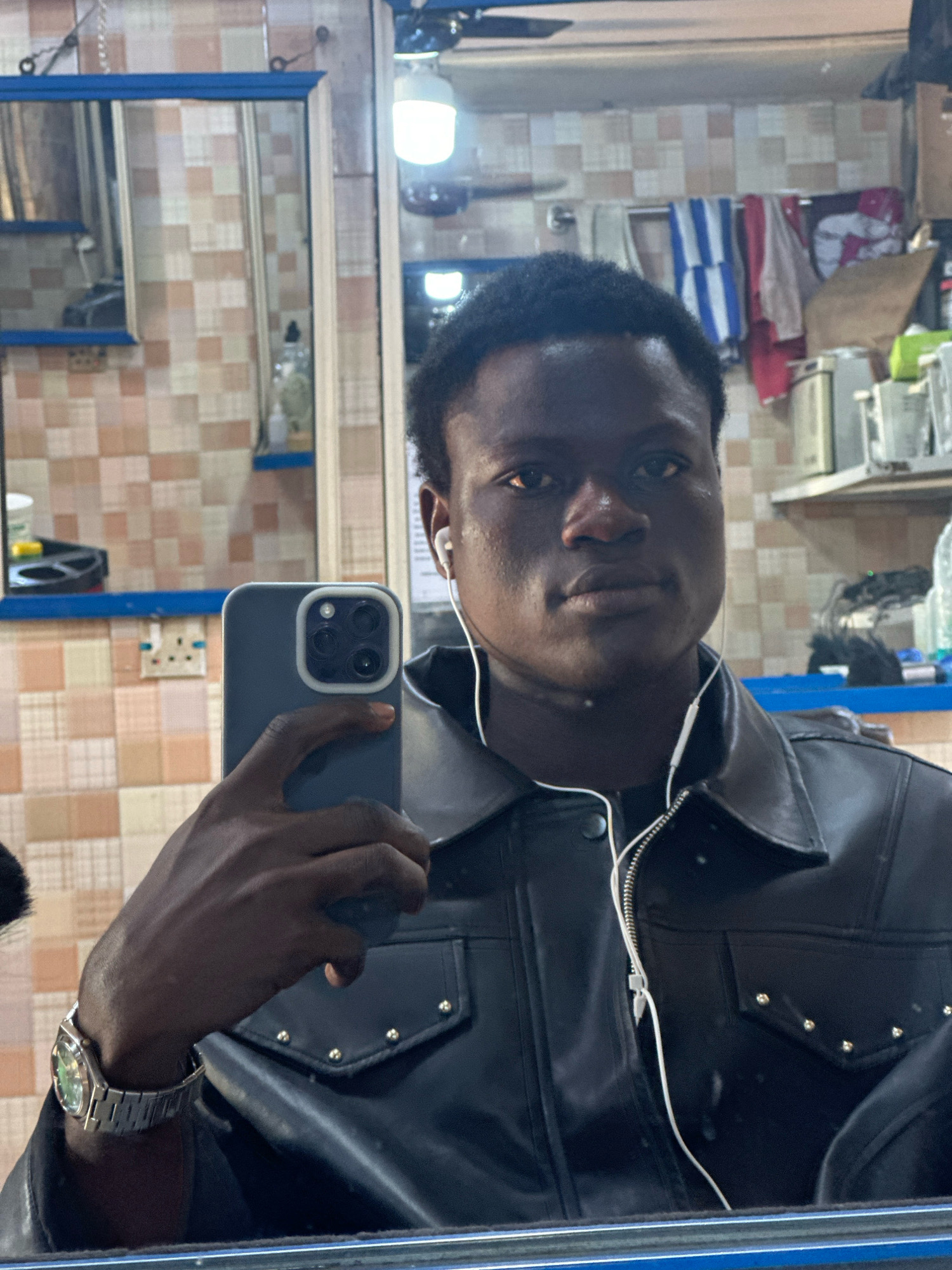 A young Black male is taking a mirror selfie in what appears to be a barbershop or salon. He is wearing a black leather jacket with silver studs on the pockets and a silver wristwatch. He has earphones in his ears and is holding a smartphone in his left hand. The background shows tiled walls, mirrors, shelving with various products, and a light fixture. The scene is brightly lit. The context of Badagry, Nigeria, suggests a commercial establishment within that city.