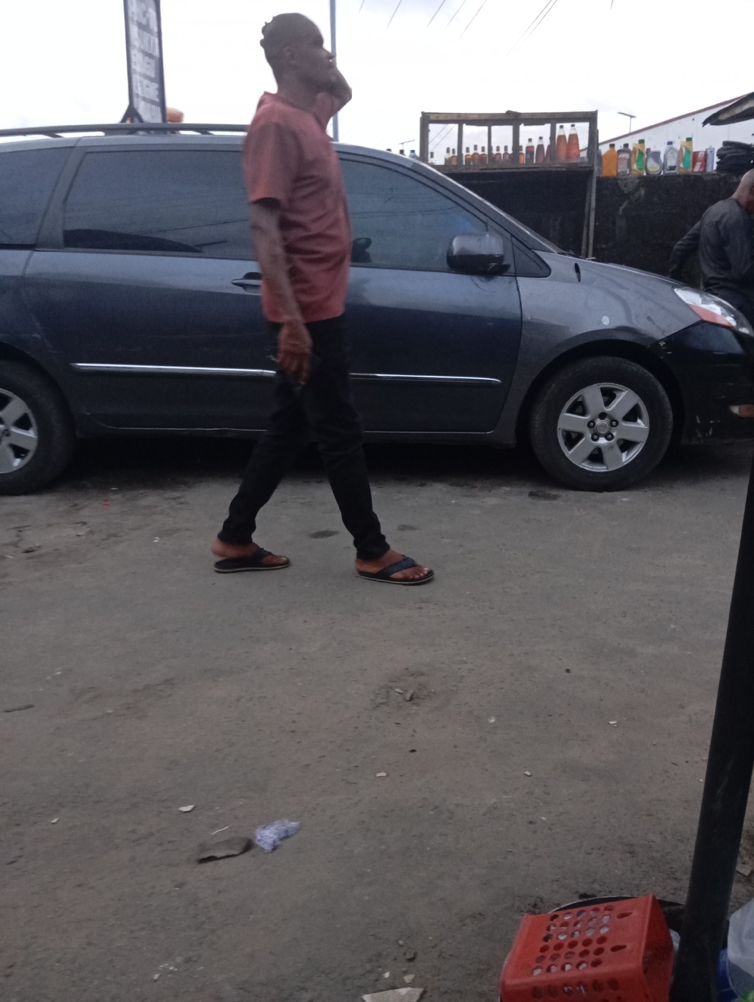 A man in a salmon-colored t-shirt and black pants walks past a dark gray minivan. He is seen in profile, with his right hand to his ear, as if on the phone. The man is wearing brown and black flip-flops. The minivan has a roof rack and is parked on a dirt or gravel road. In the background, there is a structure with shelves holding various bottles, possibly for sale. Another person, wearing dark clothing, is partially visible to the right of the minivan. The scene appears to be an outdoor market or roadside stall in Port Harcourt, Nigeria. A red plastic crate is visible in the lower right foreground. The sky is overcast.