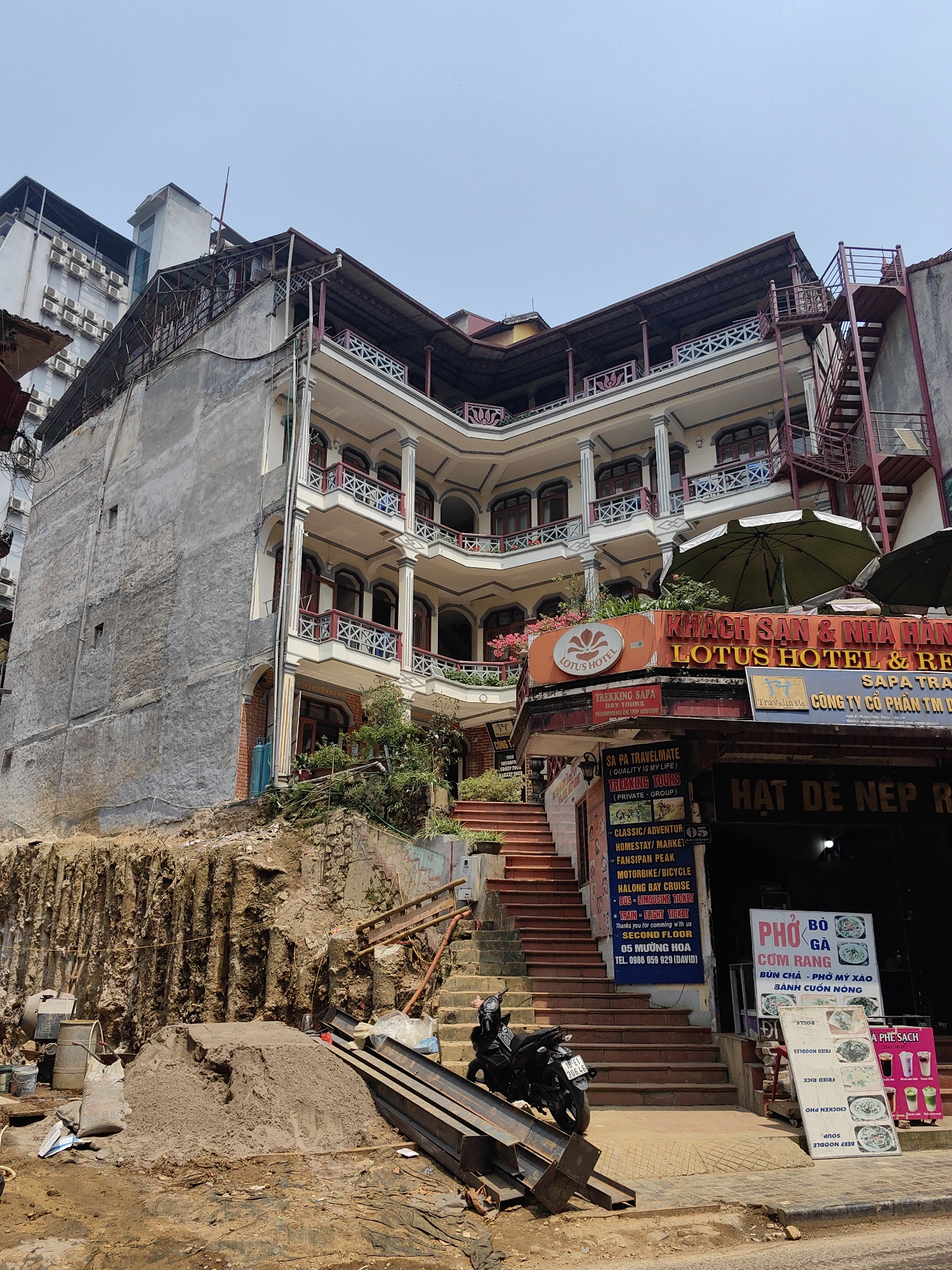 This image depicts a multi-story hotel and restaurant complex, identified by prominent signage as "Lotus Hotel & Restaurant," situated on a sloped urban street in Sa Pa, Vietnam, under a clear blue sky. The main building features a cream-colored facade with several levels of rounded balconies supported by white pillars, and dark red balustrades with decorative motifs. An external red metal fire escape staircase is visible on the right side of the building.
To the immediate left of the main structure, a raw, excavated earth wall with exposed soil and rock indicates significant ground alteration, possibly for construction or site preparation. At the base of this excavation, a large pile of sand, several sacks, and industrial equipment including a concrete mixer are present. Stacked metal beams lie on the ground adjacent to this area.
On the right, a wide set of brown-tiled steps ascends from the street level towards the hotel's entrance. A black motorcycle with license plate 24H-308.45 is parked at the base of these stairs. Adjacent to the staircase, a commercial establishment operates under a dark awning, displaying white signs advertising "PHỞ BÒ GÀ CƠM RANG" and "A PHÊ SẠCH" with various food and beverage options. Two large, dark green outdoor umbrellas are deployed in front of this establishment. A single individual, dressed in dark clothing, is partially visible ascending the steps on the far right.