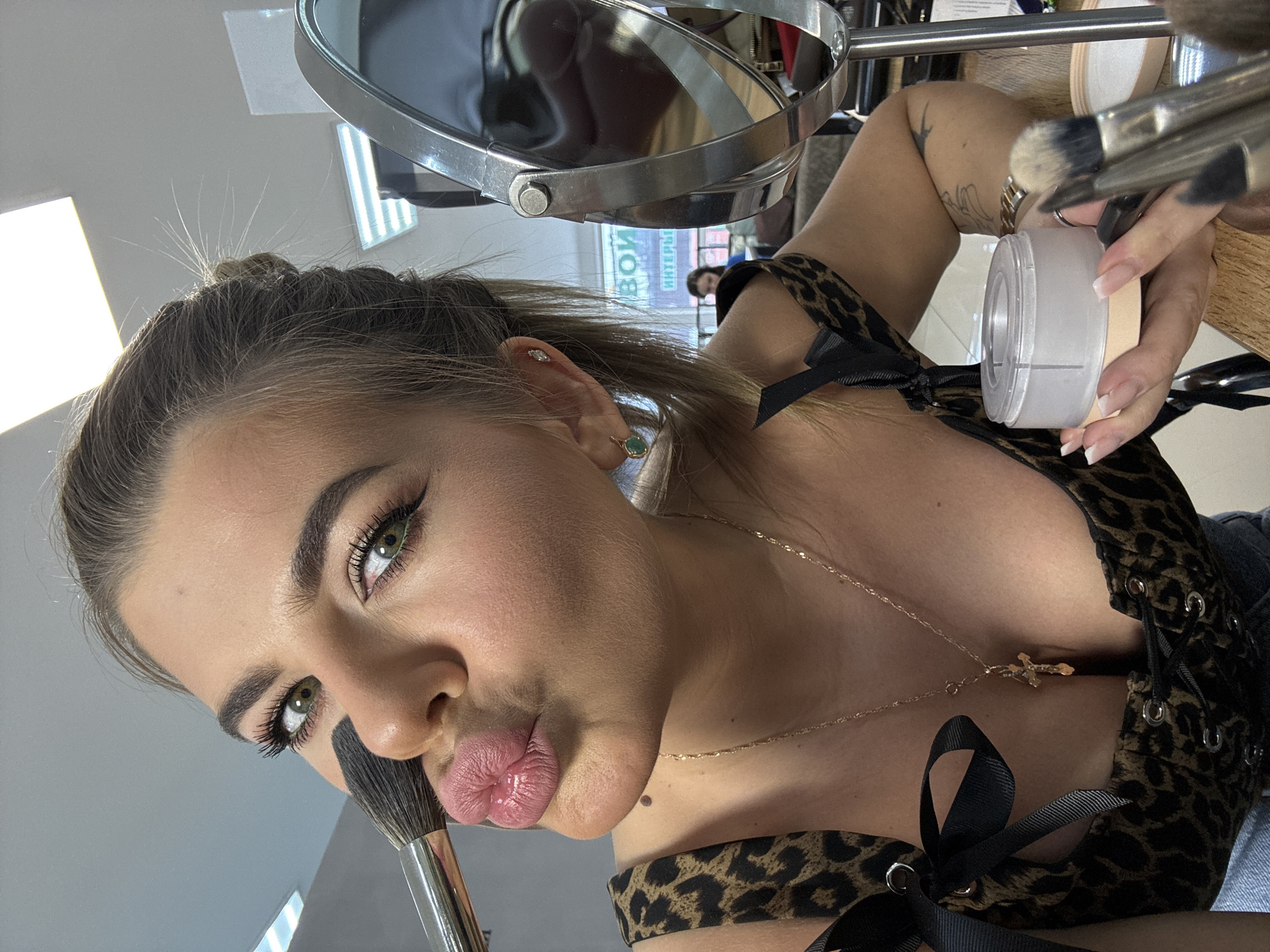 A woman poses for a close-up photograph while applying makeup. The image is taken from a low angle, looking up at the woman. She has light brown hair pulled back, green eyes with visible eyeliner, and plump lips puckered for the camera. She is holding a makeup brush to her face, and a jar of cosmetic product is visible in her hand. She wears a low-cut, leopard-print top with black ribbon details and a gold necklace with a cross pendant. A makeup mirror and additional brushes are visible in the background. The lighting is bright and appears to be indoors. In the reflected in the mirror the sign is present: “Interior in Chaikovsky”.