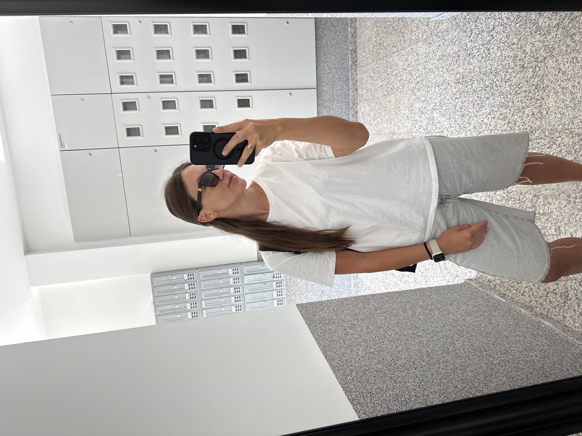 A woman is taking a mirror selfie in a well-lit, modern hallway. She is wearing a white t-shirt, light-wash denim shorts, sunglasses, and a watch on her left wrist. She is holding a black smartphone in her right hand, which is being used to take the picture. Behind her, the hallway features a speckled gray floor and a wall covered with lockers or storage units. To her left, there appears to be a textured gray wall with a diagonal white partition. The lockers in the background are arranged in a grid pattern and have small, square openings. The overall environment suggests an interior space, possibly a building with communal facilities.