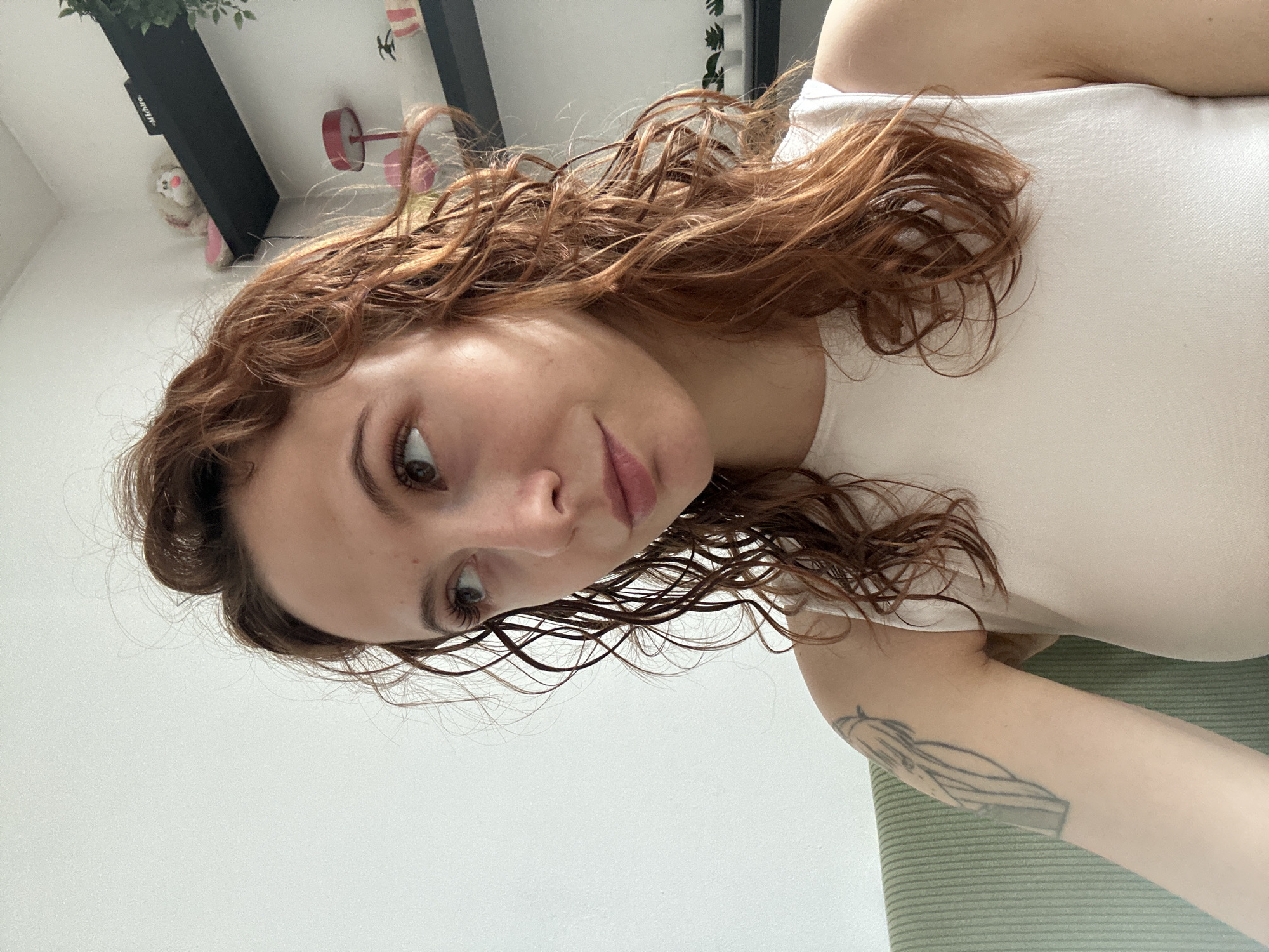 A young woman with reddish-brown, wavy, and damp hair is captured in a close-up portrait, seemingly lying down and looking upwards towards the camera. Her gaze is directed slightly to her left (our right), with a subtle, serene expression on her face. She has defined eyebrows and full lips. She is wearing a simple white sleeveless top. A black-outlined tattoo is visible on her left forearm, depicting abstract or stylized forms.
The setting appears to be indoors in Novi Sad, Serbia, with a light-colored wall or ceiling filling most of the background. In the upper left corner of the frame, elements of a room are visible, including a green leafy plant, a dark shelf, a small white fluffy toy, and a red desk lamp or decorative item. The word "ONKYO" is faintly discernible, inverted, on the black shelf. The woman is resting on a light green striped surface, possibly a mat or cushion. The bright lighting suggests it is daytime.
