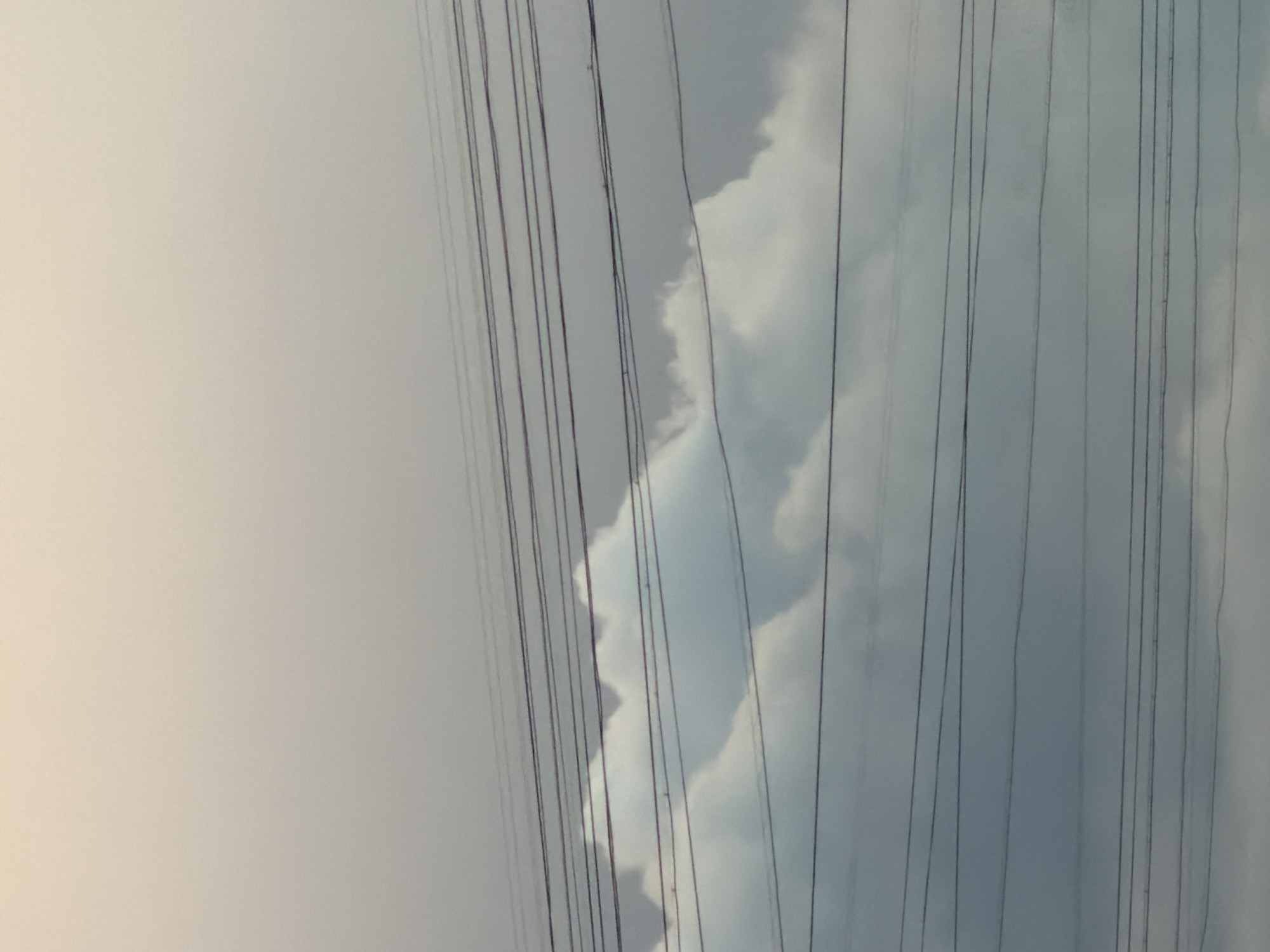 The image shows multiple parallel lines of overhead wires stretching horizontally across the frame. Behind the wires, layers of clouds are visible, with the upper clouds appearing lighter and more diffused, while the lower clouds are darker and more defined. The sky transitions from a pale, hazy tone at the top to a darker, overcast gray at the bottom. The setting appears to be outdoors, potentially in an urban or suburban area, given the presence of the power lines. This scene was captured in the city of Khimki, Russia.