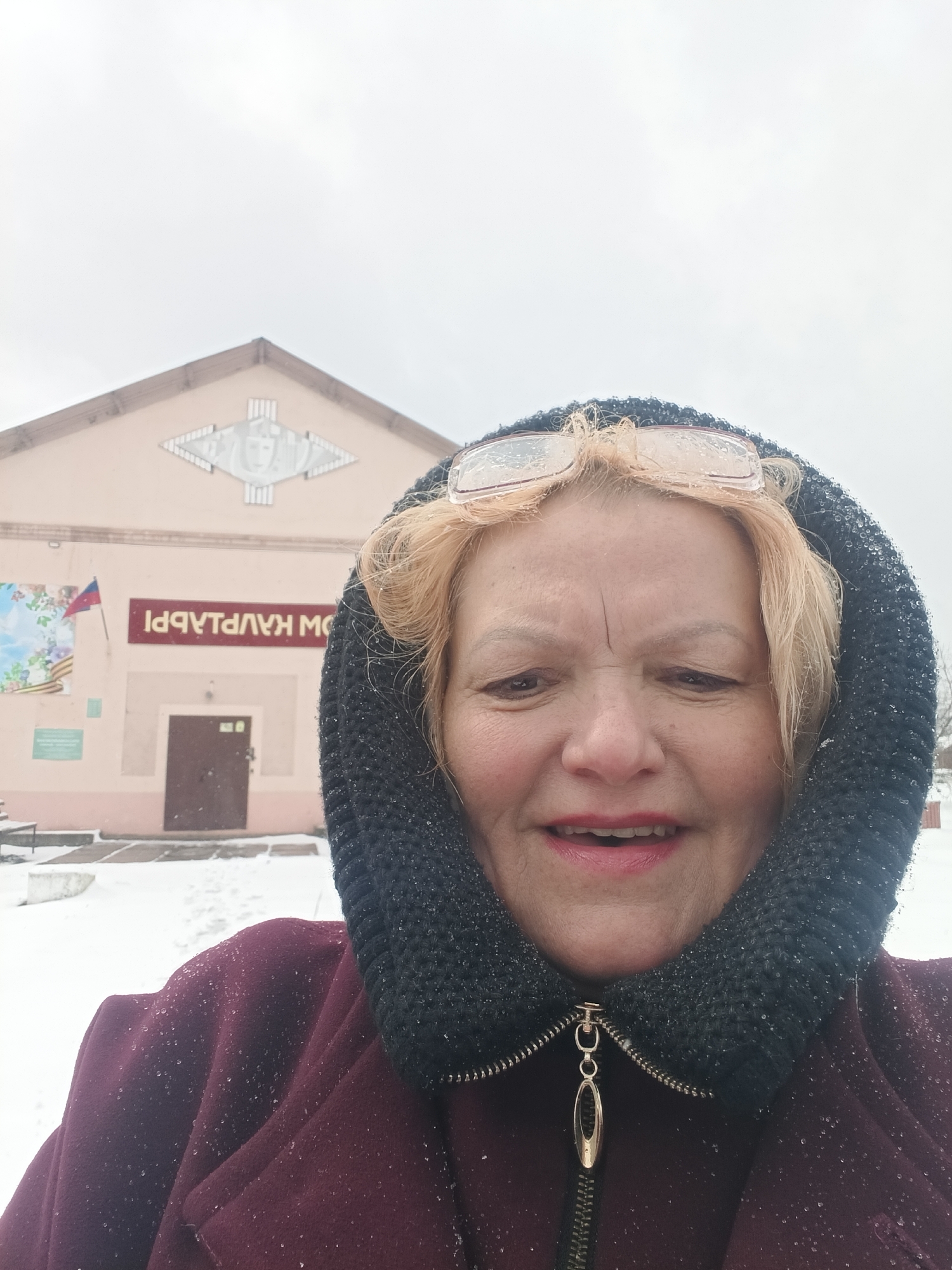 A woman takes a selfie outdoors in winter conditions, standing in front of a light peach-colored building. She has blonde hair, with her glasses pushed up onto her head, and is wearing a dark, possibly knitted, hood or cowl and a maroon-colored zippered coat. Small snowflakes or water droplets are visible on her hood, glasses, and coat, indicating cold or wet weather. She has a slight smile on her face.
The background building features a prominent decorative element on its facade, resembling a stylized theatrical mask flanked by vertical columns. Below this, there's a red sign with mirrored Cyrillic text, which, when reversed, appears to read "ДОМ КУЛЬТУРЫ" (House of Culture). A Russian flag (red, blue, white stripes, top to bottom) is also attached to the building's left side. The ground in front of the building is covered in snow, and the sky is overcast, suggesting a daytime scene. The building also has a brown door. The location is Красный Маяк, Russia.