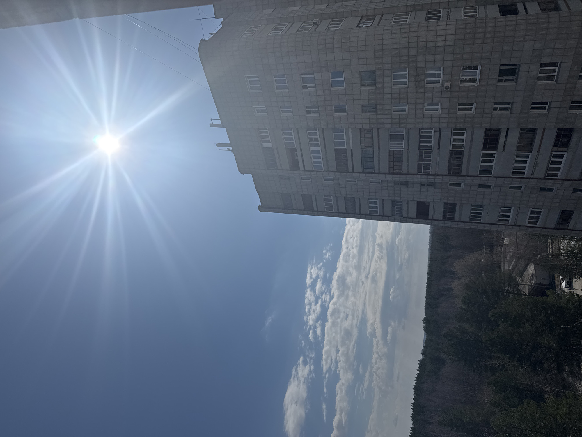 The image presents a bright, sunny outdoor scene in Chaikovsky, Russia, dominated by a large, multi-story building, a vast forest, and a vibrant sky.
On the right side of the frame, a tall, utilitarian-looking building stands, characterized by its grey, grid-patterned facade. Numerous rectangular windows, some appearing as balconies, are interspersed across its many floors. Subtle details like an external air conditioning unit on one wall and small antennas along its roofline are visible. Wires stretch across the upper portion of the sky, close to the building.
The sun is a powerful presence in the upper left of the image, emitting bright, star-like rays across a clear, light blue sky. The sky transitions to a deeper blue towards the horizon, where scattered white cumulus clouds dot the expanse above the distant landscape.
A dense, expansive forest forms a dark green band across the lower portion of the image, stretching into the background. Beyond the immediate tree line, faint structures, possibly distant buildings or industrial elements, and a small white car on a road are discernible against the horizon, suggesting the edge of a developed area blending into nature.
The time of day is clearly sunny daylight, likely morning or afternoon. The weather appears clear and pleasant. There is no visible human activity or text. The building's architecture suggests a style common in post-Soviet countries, providing a distinct cultural and historical cue for the setting.