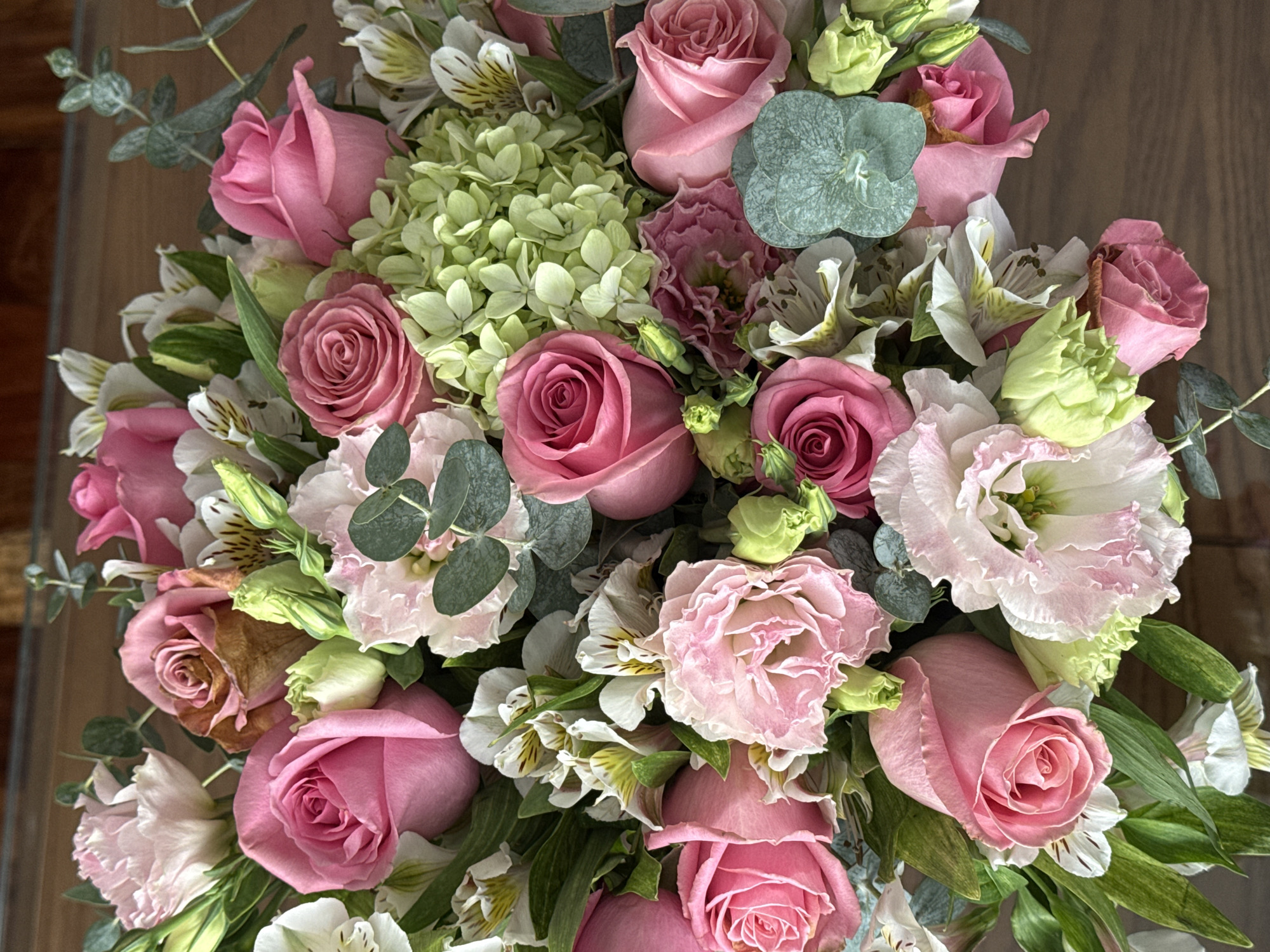 The image displays a bouquet of flowers dominated by pink roses. There are also white alstroemeria, light green hydrangeas, and pink lisianthus flowers interspersed throughout the arrangement. Green eucalyptus leaves provide additional foliage. The background is out of focus and appears to be a wooden surface.
