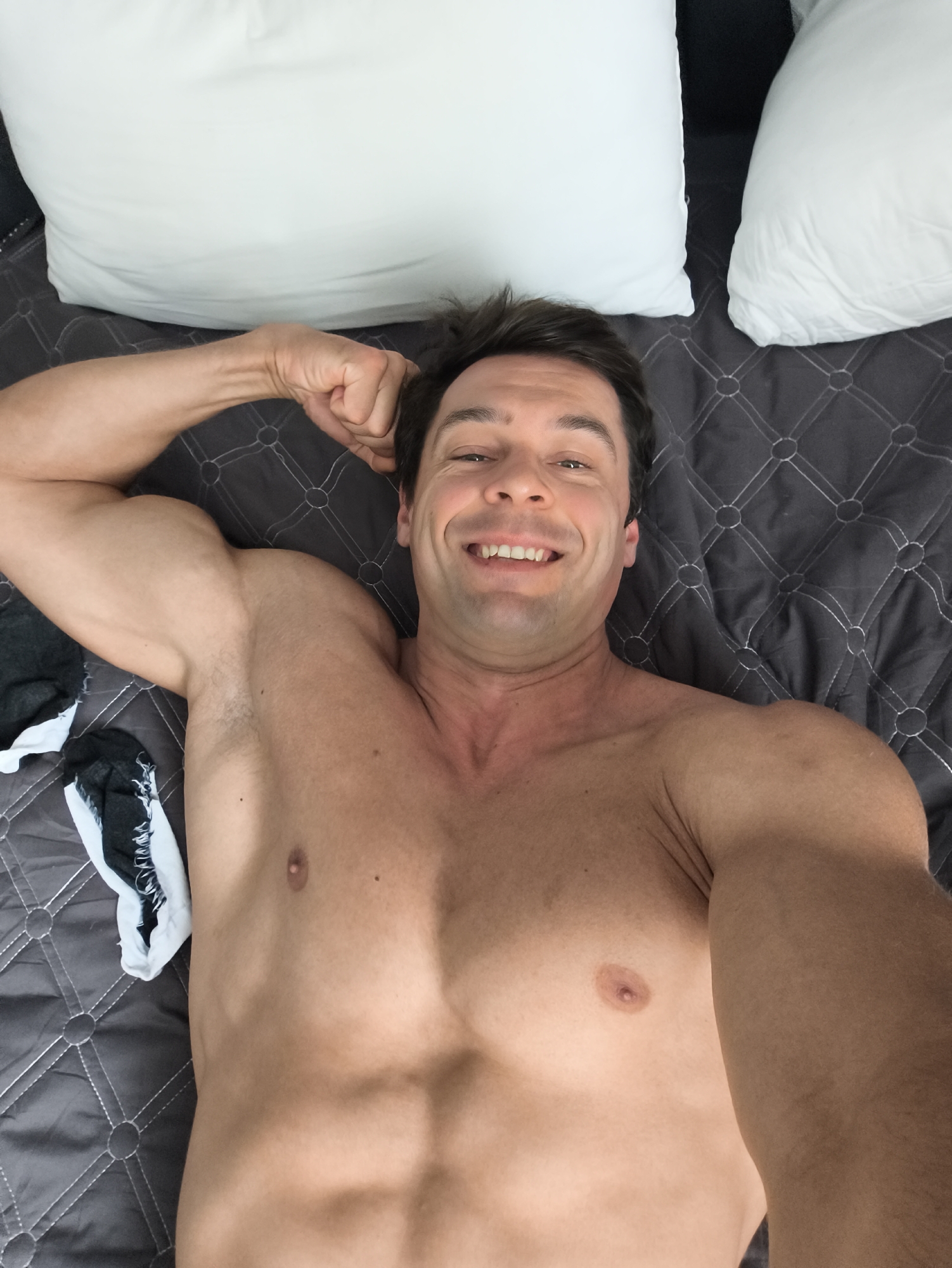 A shirtless man is lying on a gray quilted bedspread with two white pillows behind him. He is smiling and flexing his right arm, with a pair of black and white socks lying to his left. The location cannot be determined from the image.