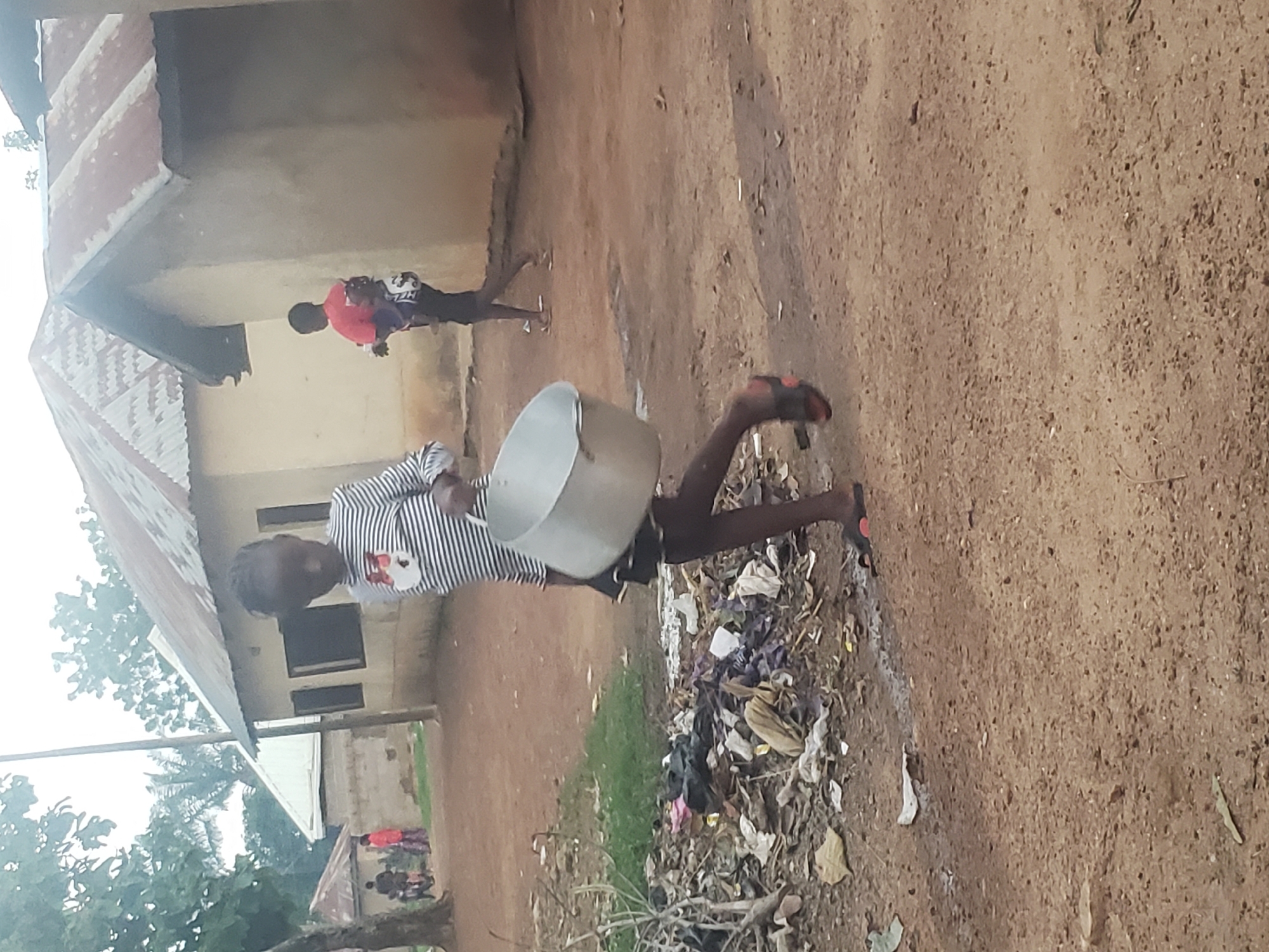 The image depicts a scene in Gumel, Nigeria, featuring a young girl walking across a dirt path while carrying a large metal pot. The girl, wearing a horizontally striped long-sleeved shirt and a dark skirt, is in motion, her body angled towards the right of the frame. She is barefoot, wearing flip-flops on her feet. To her left, a pile of assorted trash, including plastic bags and debris, lies on the ground.
Behind the girl, another individual, a person wearing a red t-shirt and dark pants, is visible in the mid-ground, appearing to walk away from the camera. They are carrying something on their back. In the background, several buildings with corrugated metal roofs are present, suggesting a residential area. The overall environment appears somewhat unkempt, with the dirt path and scattered refuse. Trees and foliage are visible in the upper left portion of the image. The sky is overcast, contributing to a diffused light.