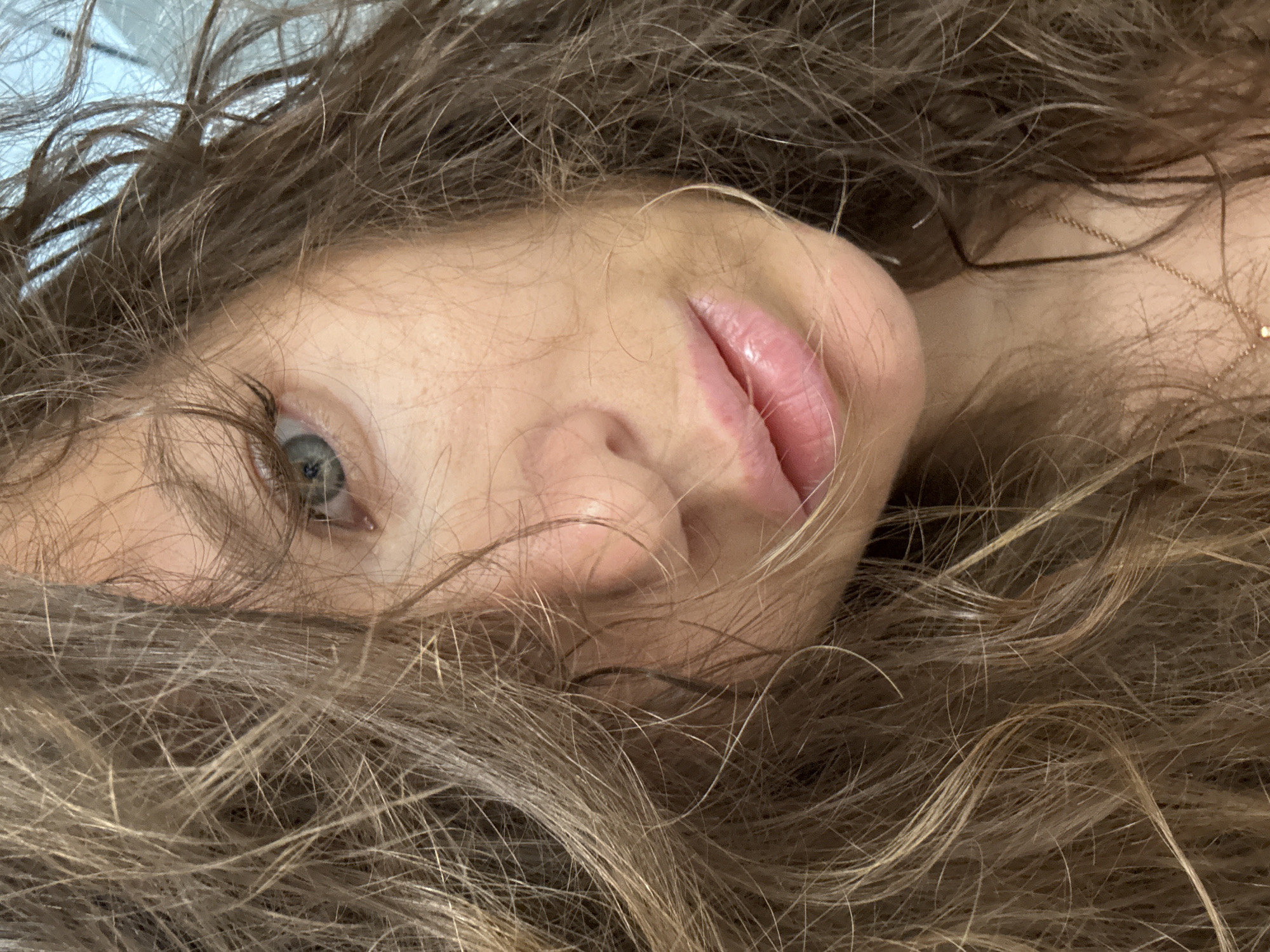 A close-up, eye-level shot captures a portion of a person's face, mostly obscured by an abundance of wavy, light brown and blonde hair. The person appears to be lying down, with their head tilted slightly to the right. The visible features include the left eye, which is a light blue-grey color, part of the nose, and the full lips, which are parted slightly and have a glossy, pink appearance. The skin tone is fair.
The mass of hair frames the face, spreading out around it. Individual strands are visible, some lighter, some darker, suggesting natural highlights. On the right side of the image, near the neck area, a delicate gold chain necklace with a small, round pendant is visible against the skin, peeking through the hair. The background is out of focus and appears to be a light, possibly blue or white, surface. The overall impression is an intimate portrait, possibly a selfie. There is no visible text. The context indicates the location is Khimki, Russia.