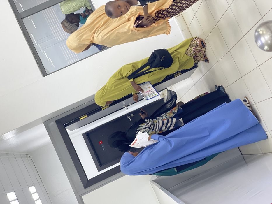 An indoor scene within a building in Zaria, Nigeria, depicts five individuals and reflections of others in what appears to be a waiting or reception area. The environment features white walls, a white tiled floor, and overhead fluorescent lighting. Two dark grey doors with silver handles are visible on the left, one partially open, revealing a darker interior. The visible door displays a small red square emblem and faint text "Opening Hr."
From left to right:
1. A woman, obscured from the waist down, wears a blue abaya and hijab, holding white papers in her hands.
2. Next to her, a woman in a black abaya and hijab, with a striped and floral garment visible underneath, holds a mobile phone in her left hand and papers in her right. She faces the central door.
3. A woman in a lime green abaya and hijab, carrying a black shoulder bag, extends her right hand towards the door frame or a wall-mounted device next to the door. She holds a document with a colorful emblem in her left hand.
4. On the right, a woman in a light orange/peach abaya and hijab stands, holding the hand of a young child.
5. The child, partially visible in the lower right foreground, has short dark hair and wears a dark top, looking directly towards the camera.
A horizontal window on the right wall reflects other individuals, including an adult with a patterned head covering and a child, suggesting additional people are present outside the immediate frame. A metallic spherical object is visible on the floor in the bottom right corner, possibly part of a barrier or fixture.