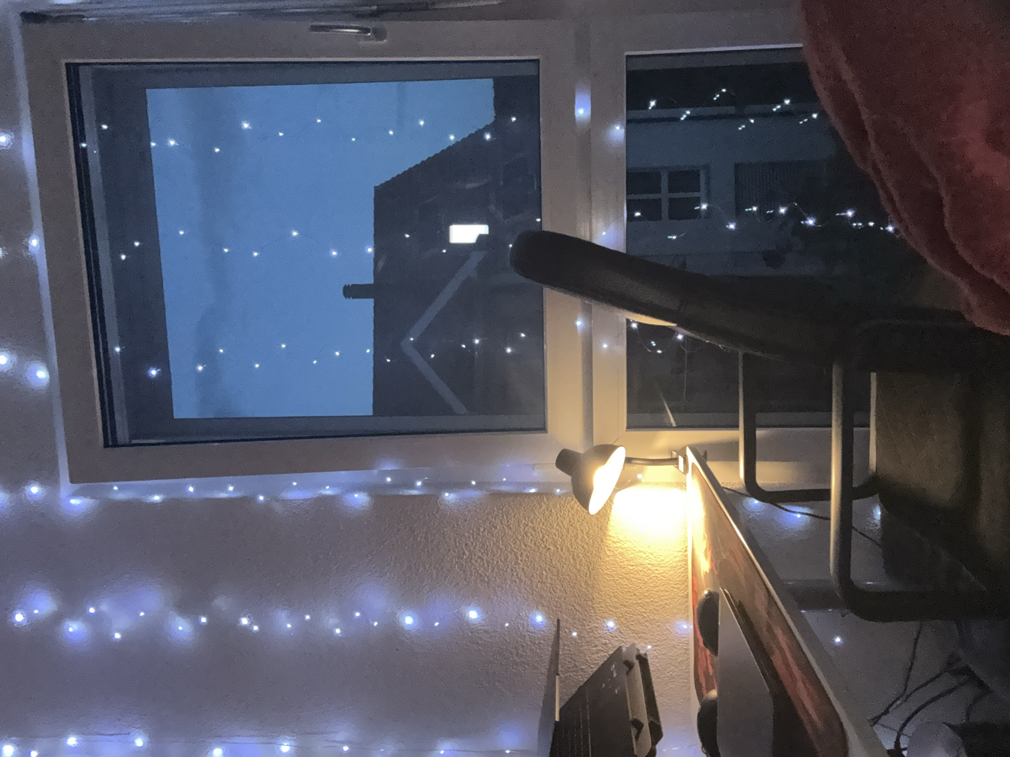 A nighttime interior scene is depicted, likely in Lufingen, Switzerland, with a desk, a chair, a window, and decorative lights. The desk, made of light-colored wood with a dark, patterned mat, holds a laptop and a mouse. To the left of the desk, a black desk lamp with a metallic finish is illuminated, casting a warm yellow glow. The wall behind the desk is textured and adorned with strings of small, blue-white fairy lights.
A dark-colored office chair with a high back is positioned in front of the desk. The scene is dominated by a large window that looks out onto a twilight sky. The sky is a deep blue, dotted with numerous small white lights, some of which appear to be part of the same fairy light string as those indoors, while others might be distant lights or stars. Reflected in the window are silhouettes of a building with a pitched roof and a chimney, and another building with multiple windows.
The overall ambiance is cozy and intimate, suggested by the soft lighting and the decorative string lights. The presence of a laptop and desk lamp implies a workspace, possibly a home office or study area. The view from the window, with the hints of buildings and the twilight sky, grounds the scene in a specific time and place, though the precise location beyond Lufingen, Switzerland is not further specified by the visual elements.