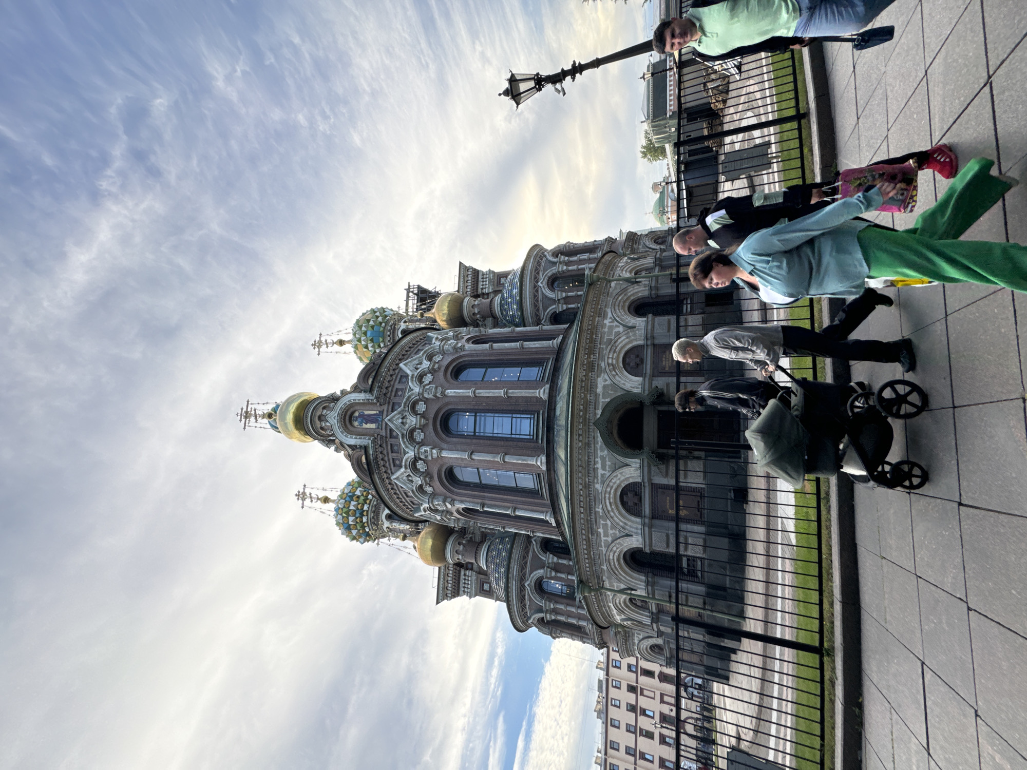 The image shows a street scene in Saint Petersburg, Russia, with the Church of the Savior on Spilled Blood prominently featured. Several people are walking along the sidewalk, some are pushing a stroller, and one person is carrying a pink bag. The sky is partly cloudy with wispy clouds, suggesting it's daytime, possibly late afternoon given the lighting. The architecture of the church is ornate, with multiple domes, some of which are gold and others are colorful and patterned. A lamppost stands on the sidewalk near the church. The people are dressed casually, and their expressions are neutral. The overall atmosphere is that of a normal day in a tourist area. There is no visible text in the image.