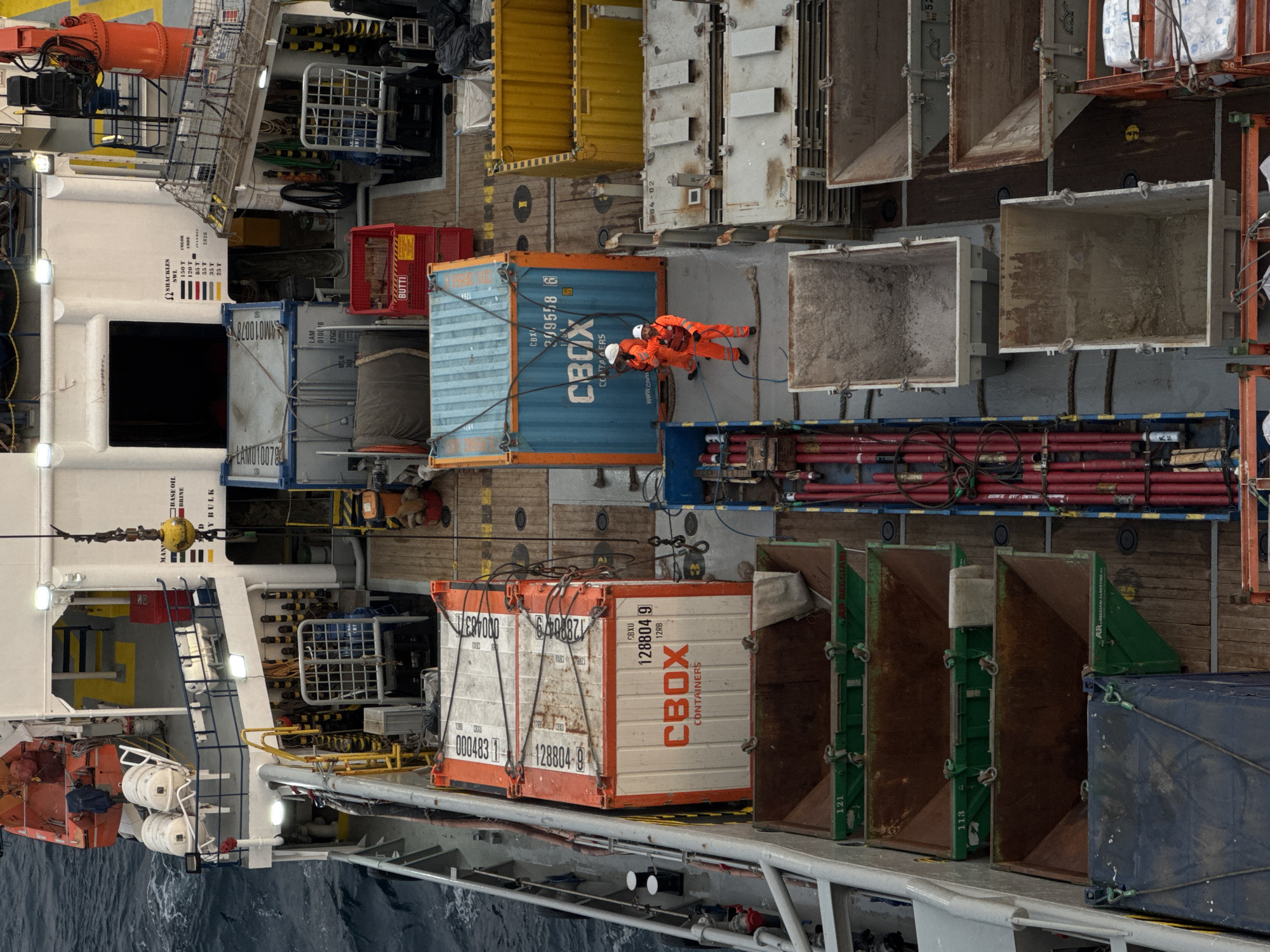 The image shows an overhead view of a ship's deck loaded with various containers and equipment. In the foreground, several large, empty, open-top bins are stacked. To the right of these bins are stacks of flat, rectangular containers with metal plating. Further to the right, there are yellow containers. In the middle ground, two individuals wearing orange safety suits and hard hats are standing near a blue shipping container. One of the individuals appears to be holding a rope. Next to the blue container is a red bin with the word "BUTTI" visible. Above this, there are orange and white shipping containers stacked. In the background, part of the ship's superstructure is visible, including lifeboats, railings, and various pieces of equipment. The deck is made of wooden planks and metal surfaces. The sea is visible on the left side of the image, with choppy water. The overall impression is of an industrial setting on a vessel, possibly engaged in cargo operations. There is no indication of a specific location within a city of Italy; the scene is on a ship at sea.