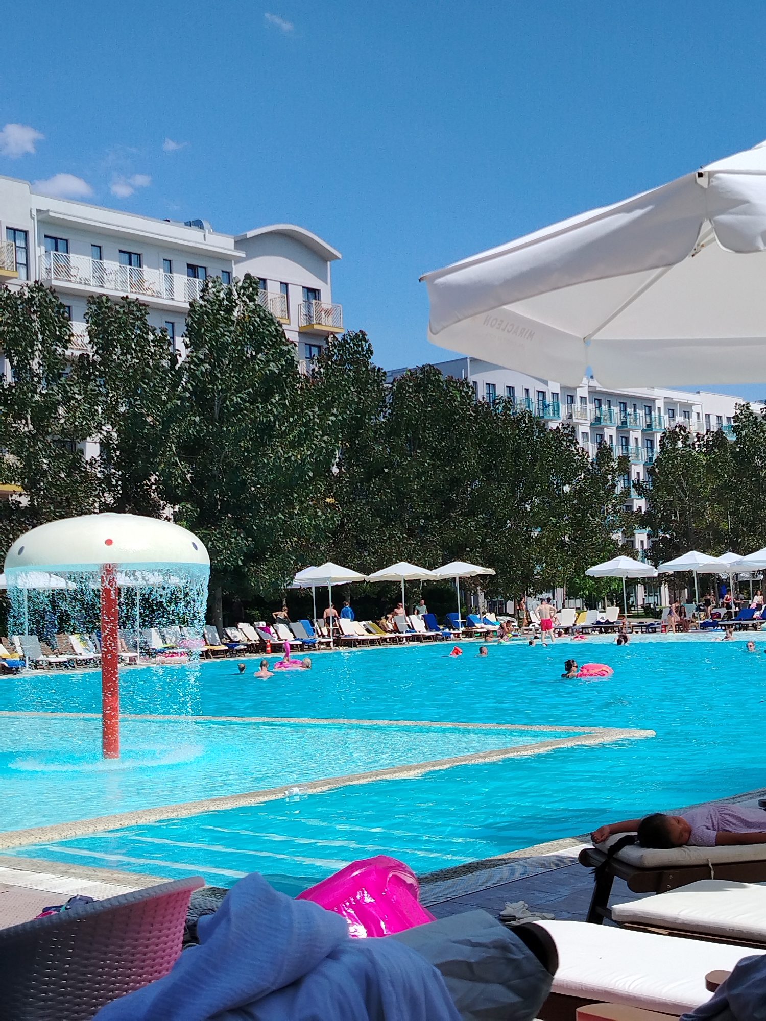 In an outdoor swimming pool complex, the scene captures a bright, sunny day with a clear blue sky. In the foreground, a large, white umbrella shades part of the view, with the word "MIRACLEGOM" visible on its surface, suggesting it might be part of a resort or hotel. The pool itself is a vibrant turquoise, with several people swimming and relaxing. Some are using inflatable toys, including a pink flamingo. A water feature resembling a mushroom with water spraying from it is positioned in the shallower section of the pool. Beyond the pool, a line of lounge chairs is set up, some occupied by sunbathers. Behind the lounge chairs, a row of lush green trees forms a backdrop, and further beyond are multi-story white buildings with balconies. The architecture of the buildings suggests a modern hotel or resort complex. In the very foreground, partially visible, are lounge chairs and towels, indicating that the viewer is also a guest enjoying the amenities.