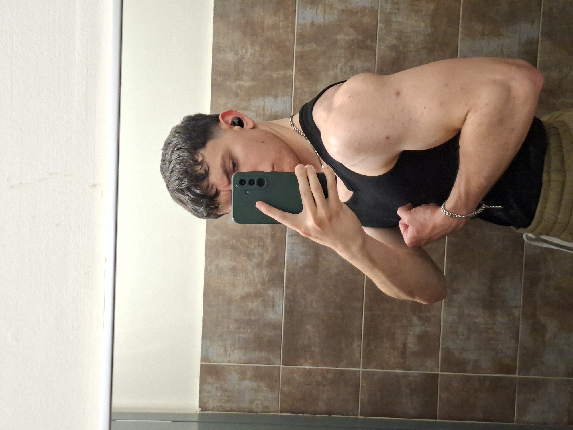 A young man is taking a selfie in a bathroom. He is wearing a black tank top and tan sweatpants with white stripes down the side. He is flexing his bicep. He has a silver chain necklace and a silver bracelet on. He is holding a green smartphone to take the picture. He is wearing an earbud in his left ear. The background consists of brown and tan tiles on the wall behind him and a white wall to his left. There is no evidence indicating the location is Plovdiv, Bulgaria.